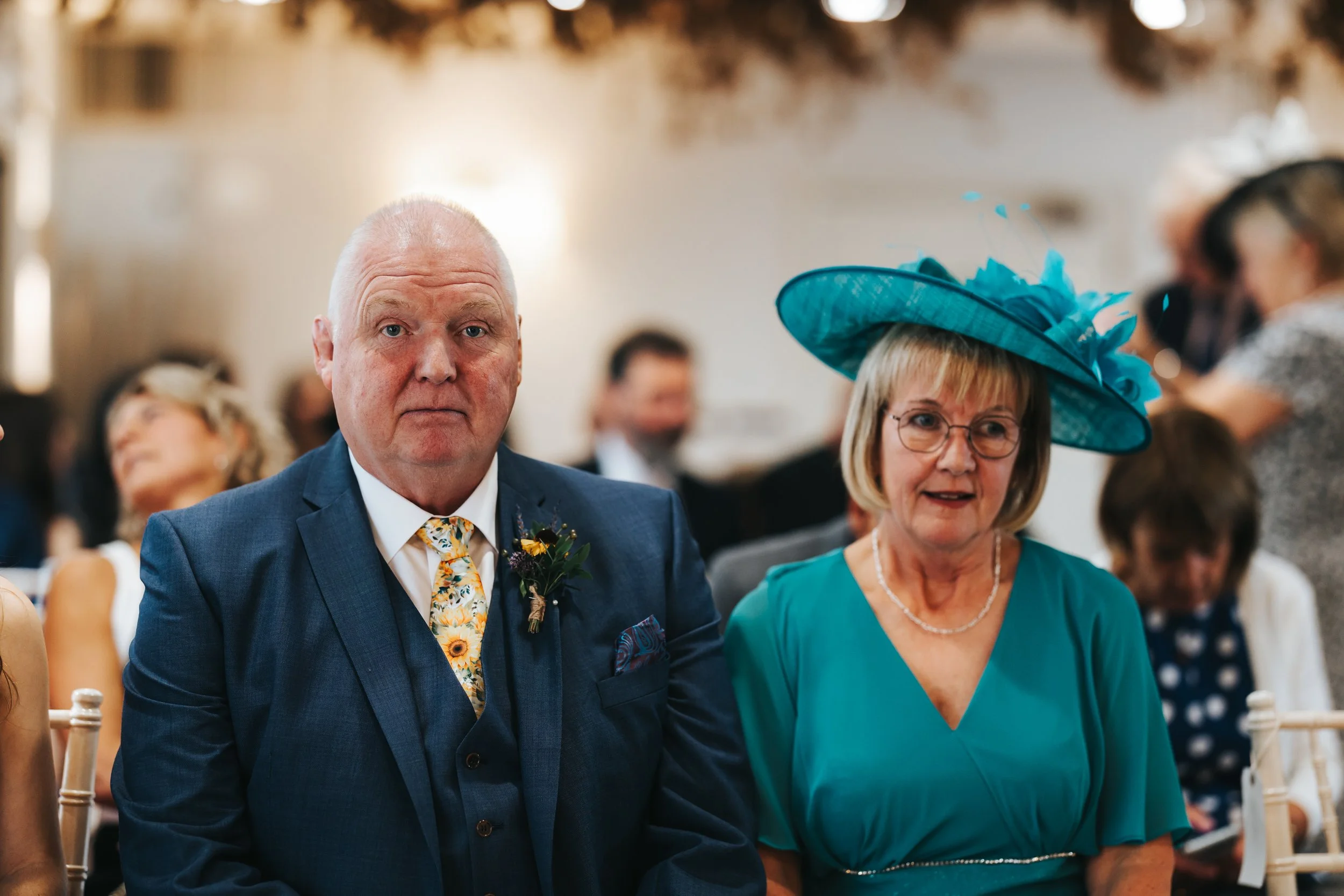 An elderly man in a blue suit with a floral tie and an elderly woman in a teal dress with a large teal hat, sitting at a formal event.