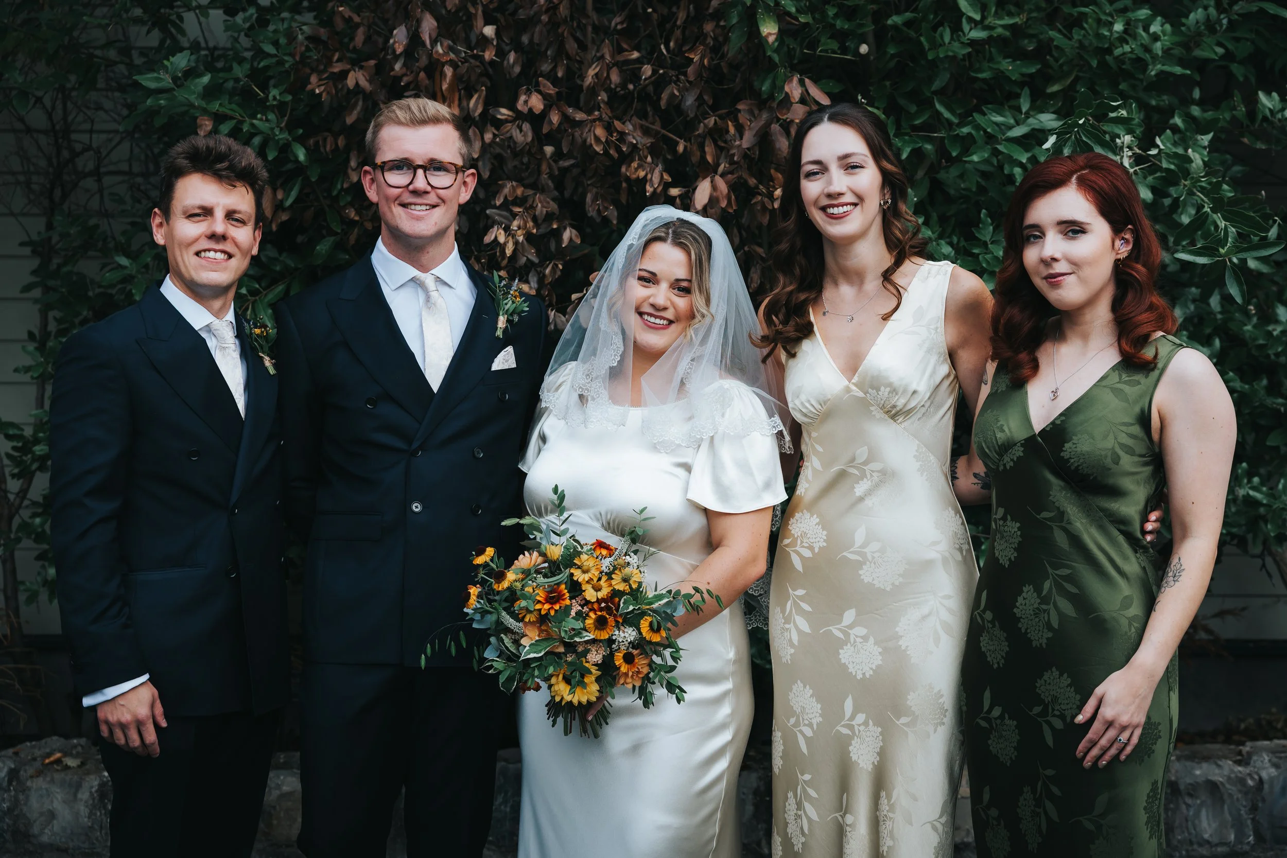 Group of six people at a wedding, with a bride holding a bouquet of flowers, standing with bridesmaids and groomsmen in front of greenery.