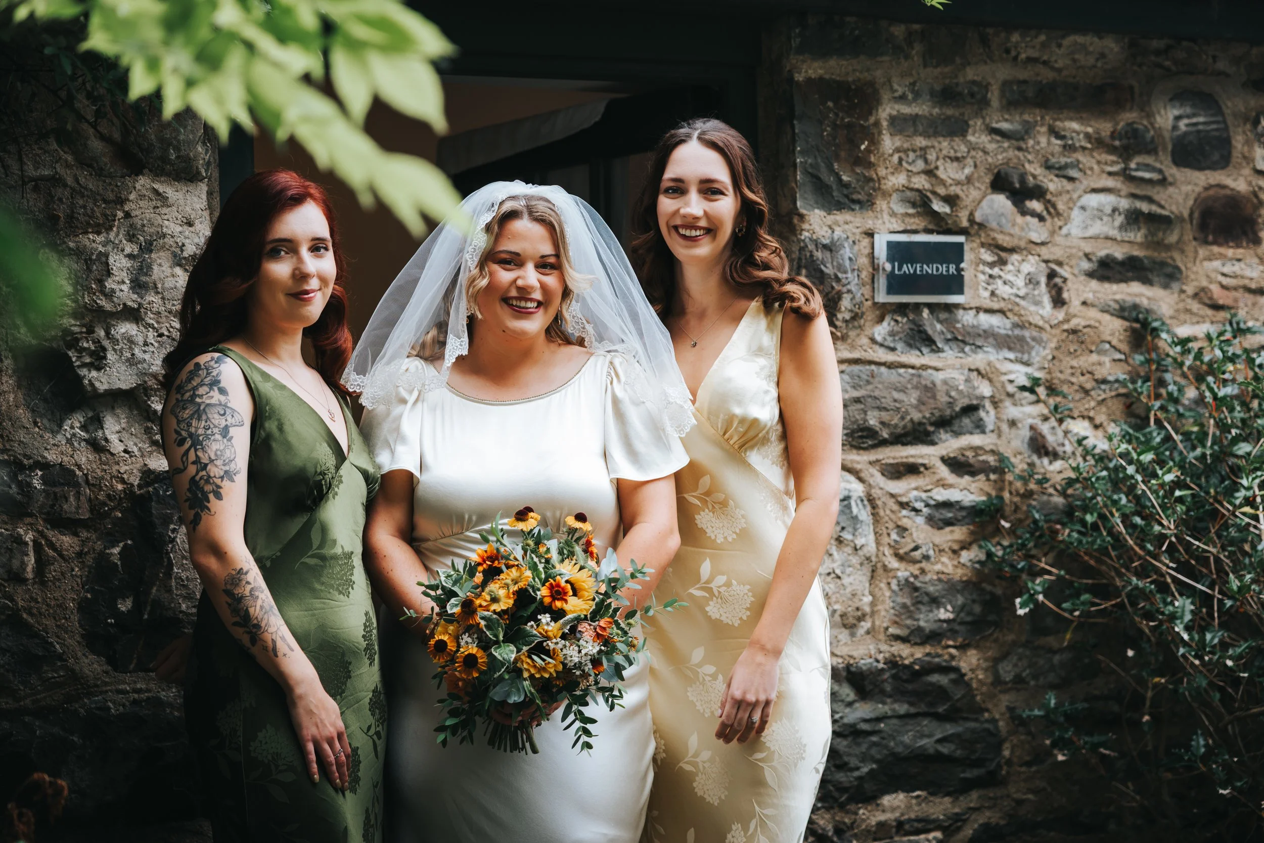Three women standing outdoors against a stone wall, with one woman in a wedding dress holding a bouquet of yellow and orange flowers, and the other two women in dresses, smiling.