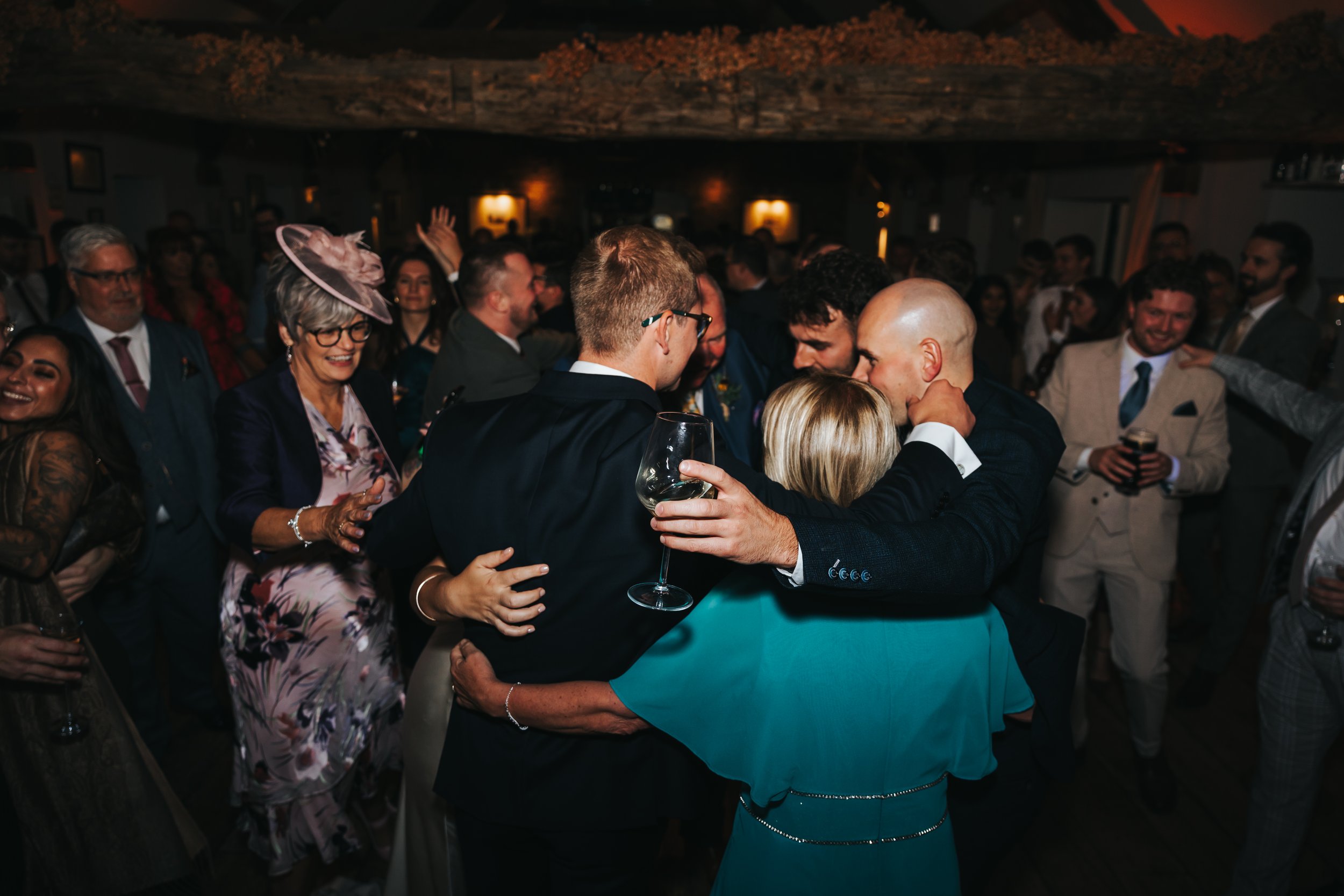 Group of people dancing and celebrating at a wedding reception, with some holding wine glasses and others embracing or smiling.
