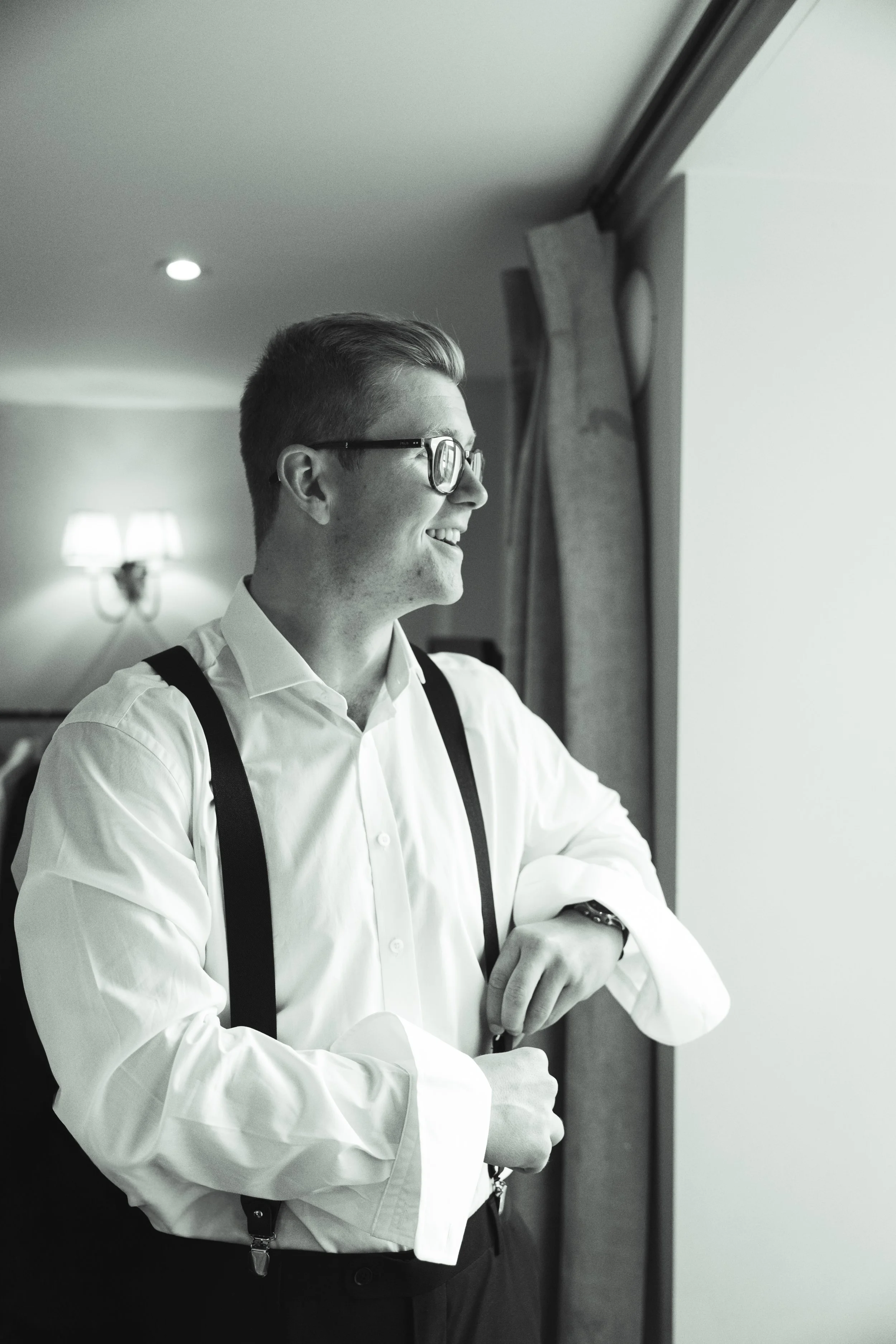 A young man in a white shirt and suspenders looking out a window with a smile, in black and white.