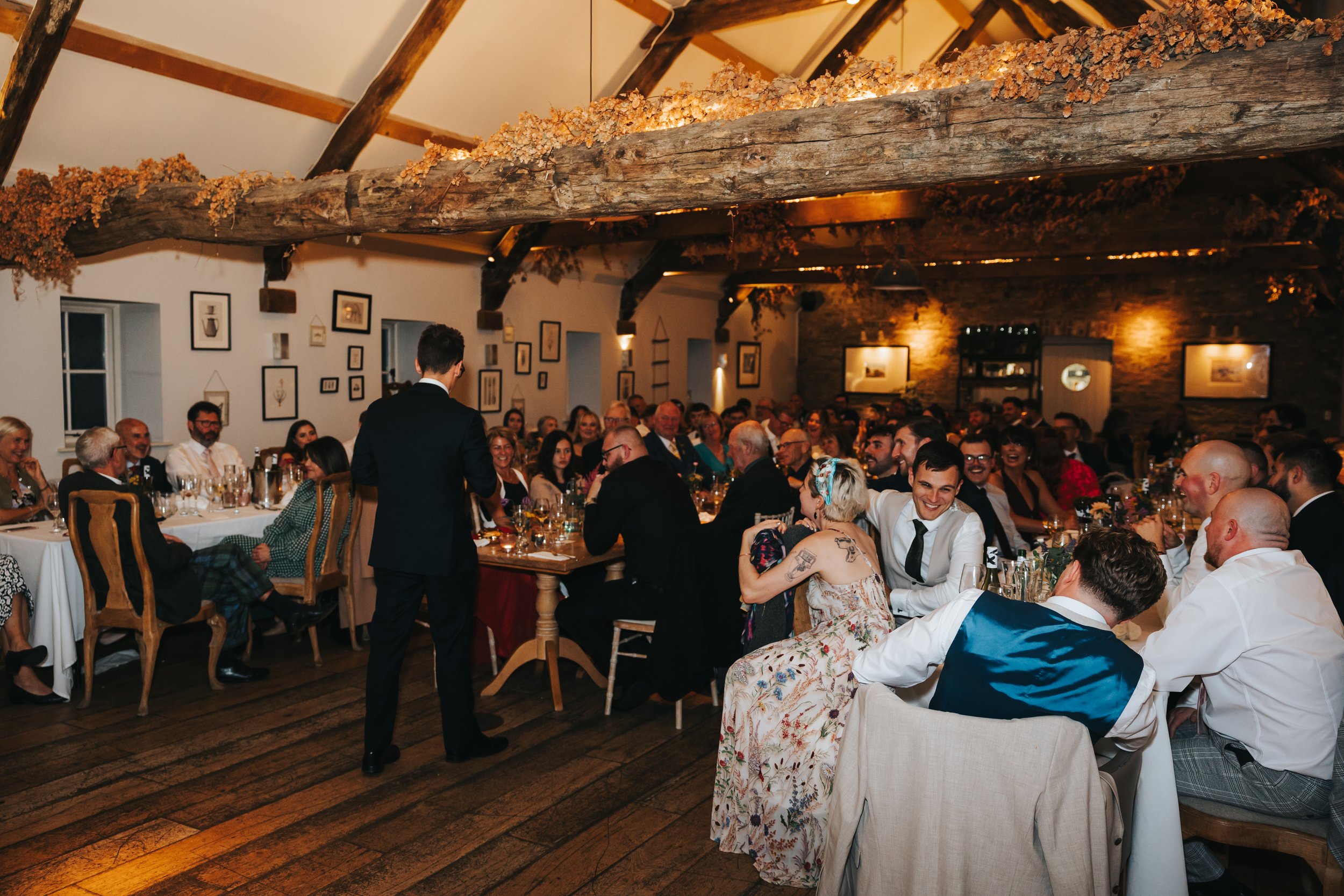 A large group of people, dressed in formal attire, seated at long banquet tables in a rustic event space with wooden beams and decorated with dried flowers hanging from the ceiling, attending a celebration or wedding reception.