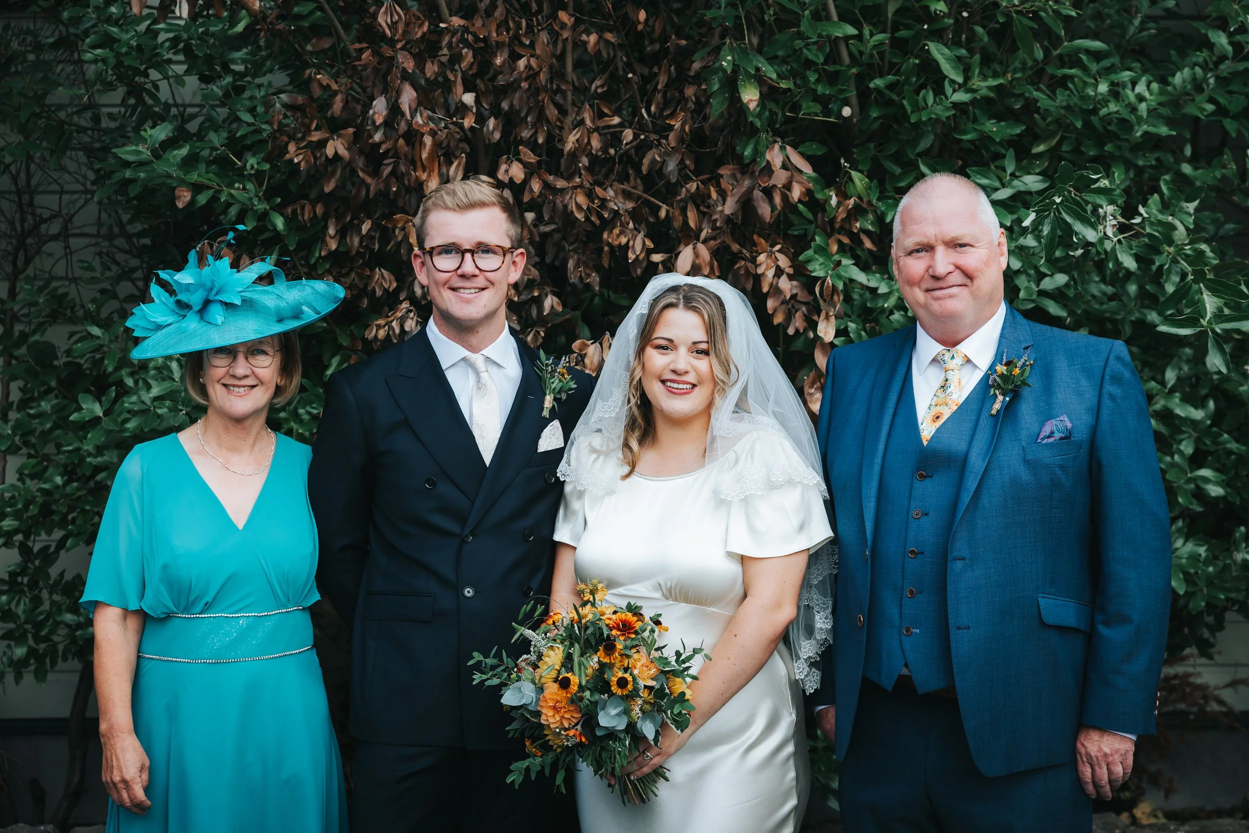 A group of five people, including a bride and groom, standing outdoors in front of green and brown foliage, smiling at the camera.