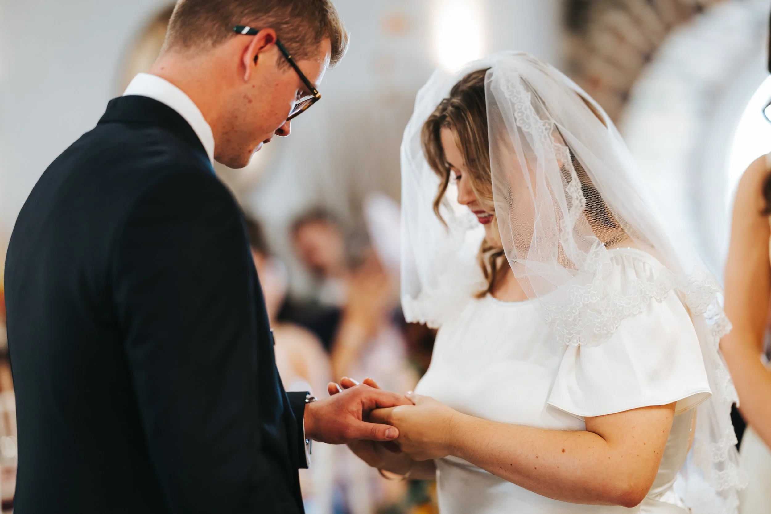 A bride and groom at a wedding ceremony holding hands and looking at each other inside a venue with blurred guests in the background.