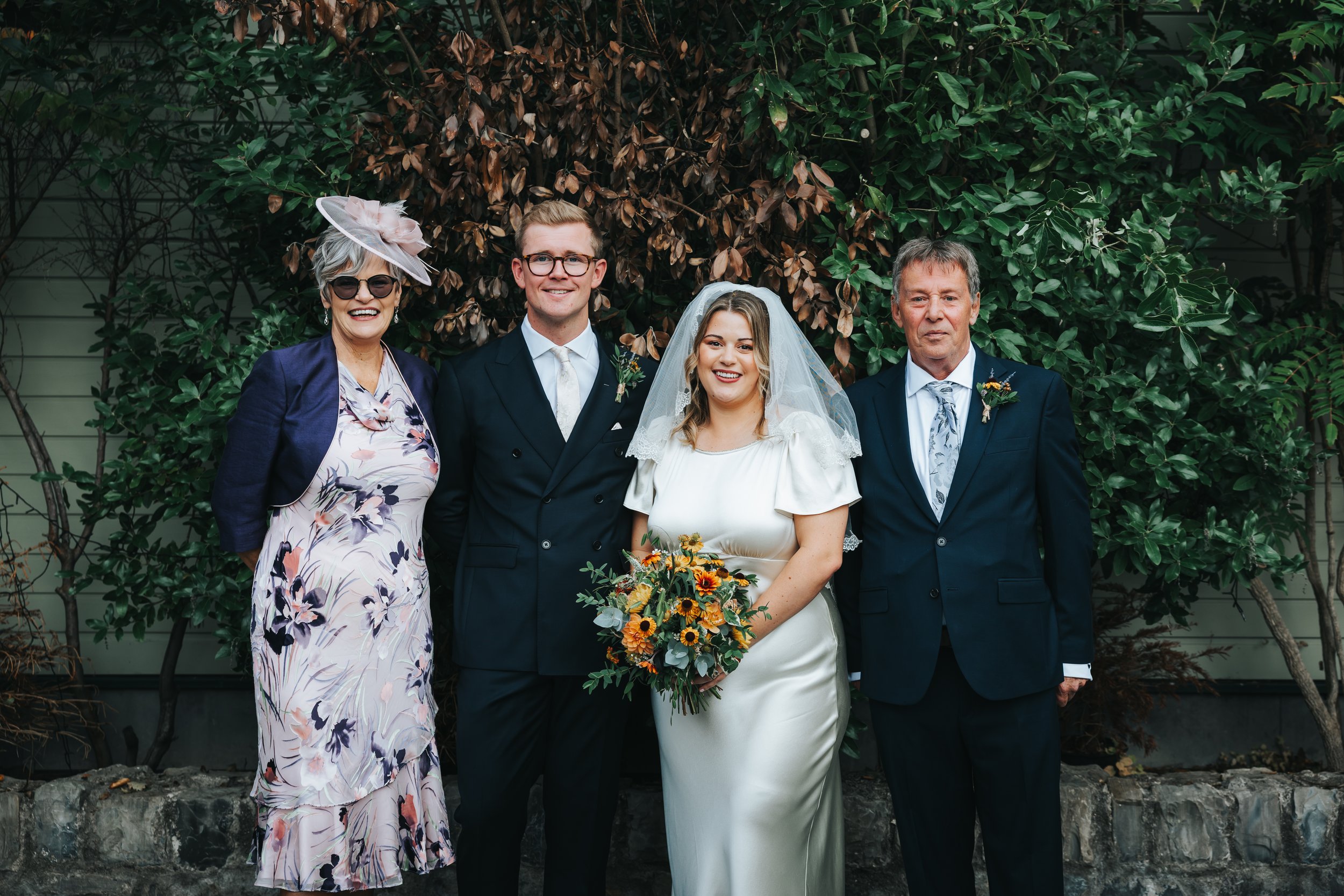 A group of five people at a wedding standing outdoors in front of green foliage. From left to right: an older woman wearing a floral dress, navy blazer, and a large pink hat with sunglasses; a young man in a navy suit, white shirt, and glasses; a bri