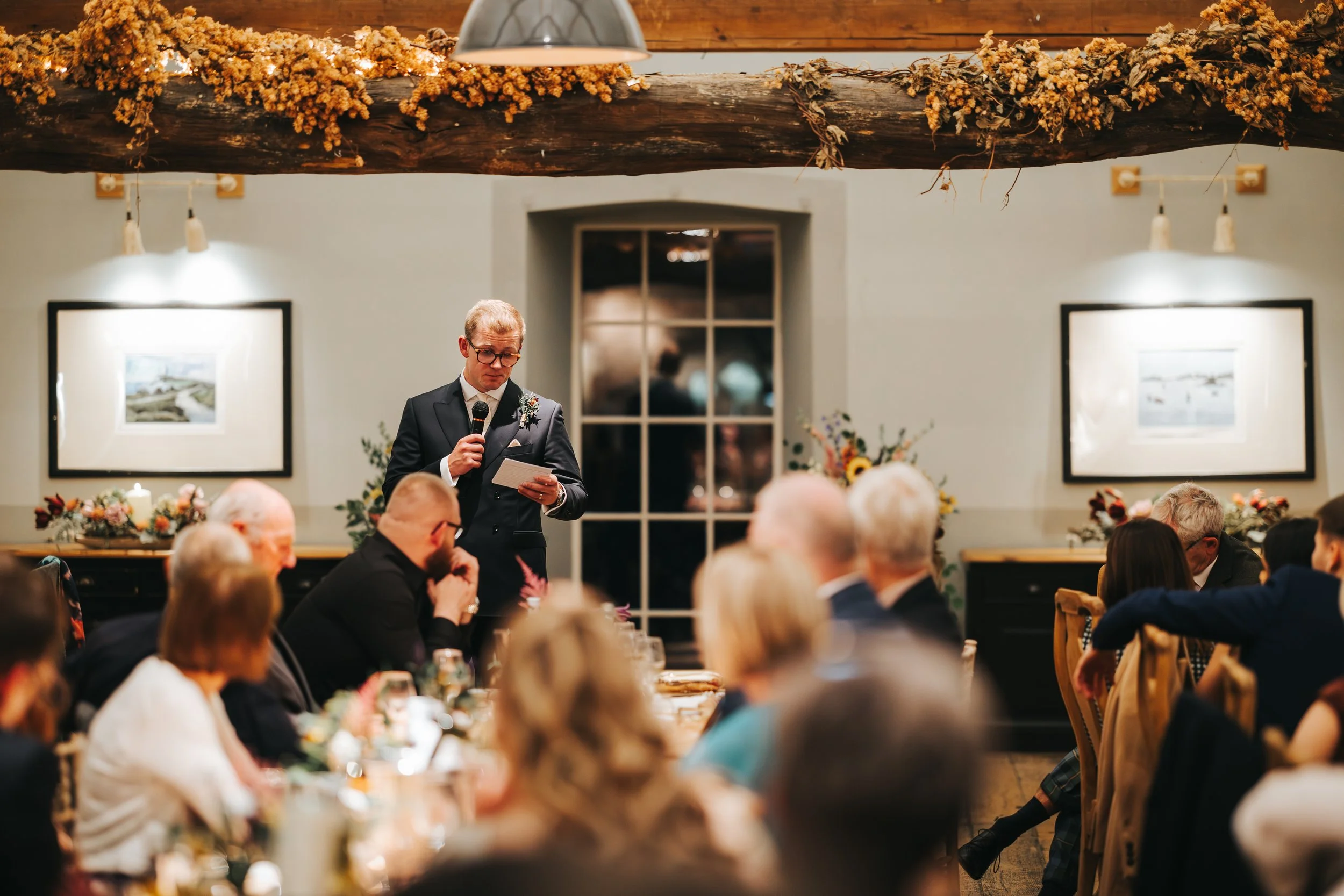 A man in a suit giving a speech at a wedding reception, holding a microphone and reading from a paper, surrounded by seated guests in a decorated indoor venue.