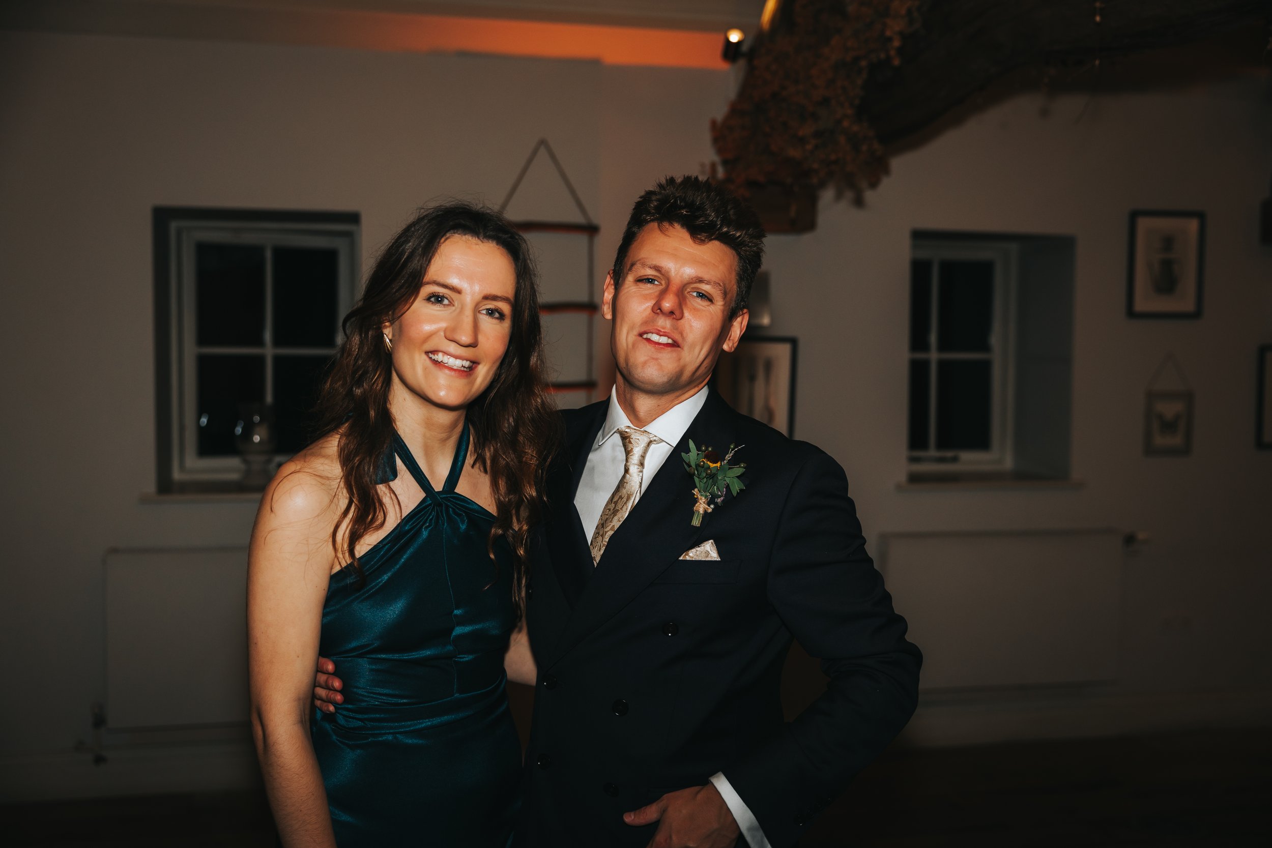 A smiling woman in a teal dress standing next to a man in a dark suit with a boutonniere, in a warmly lit room with framed pictures on the wall.