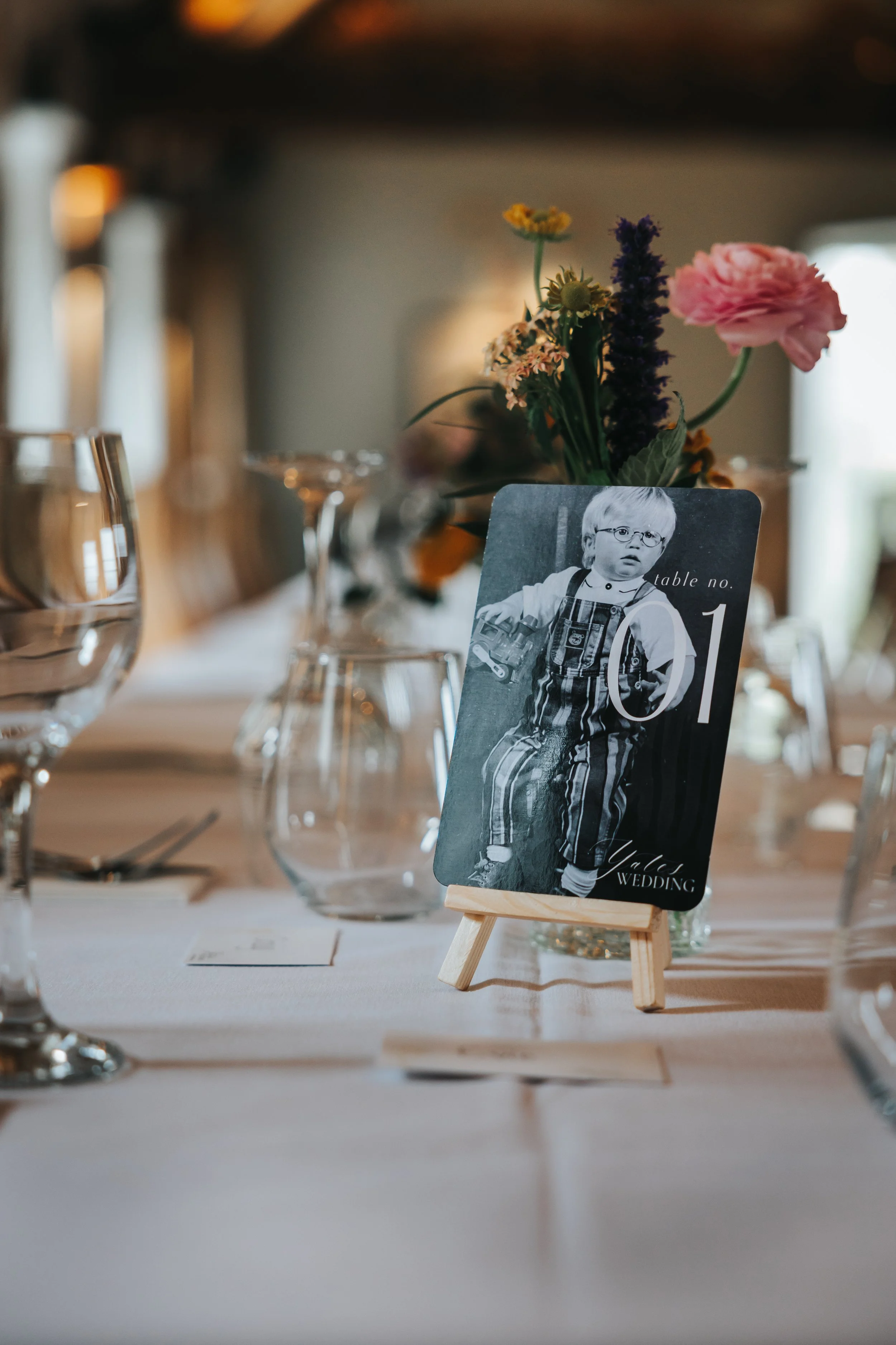 A wedding reception table setting with a black-and-white photograph of a young boy in striped overalls as a table number card, surrounded by wine glasses, floral centerpiece, and tableware.