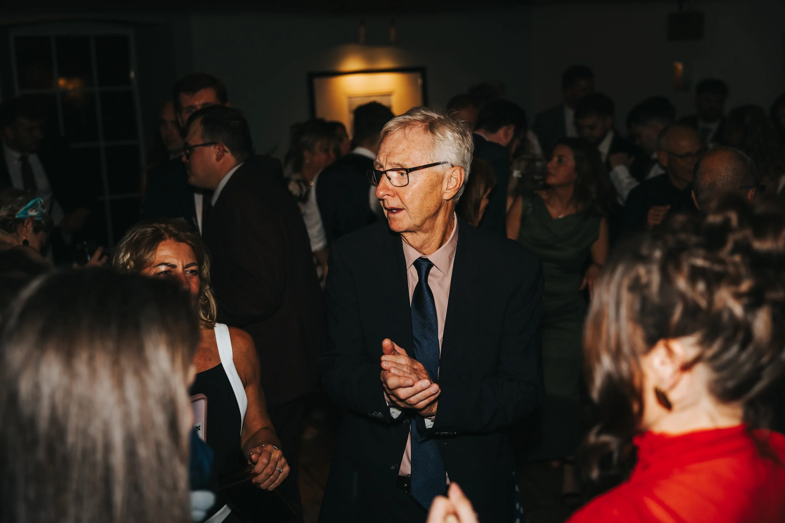 An older man in glasses, a black suit, white shirt, and blue tie, clapping while talking to a woman at a crowded event.