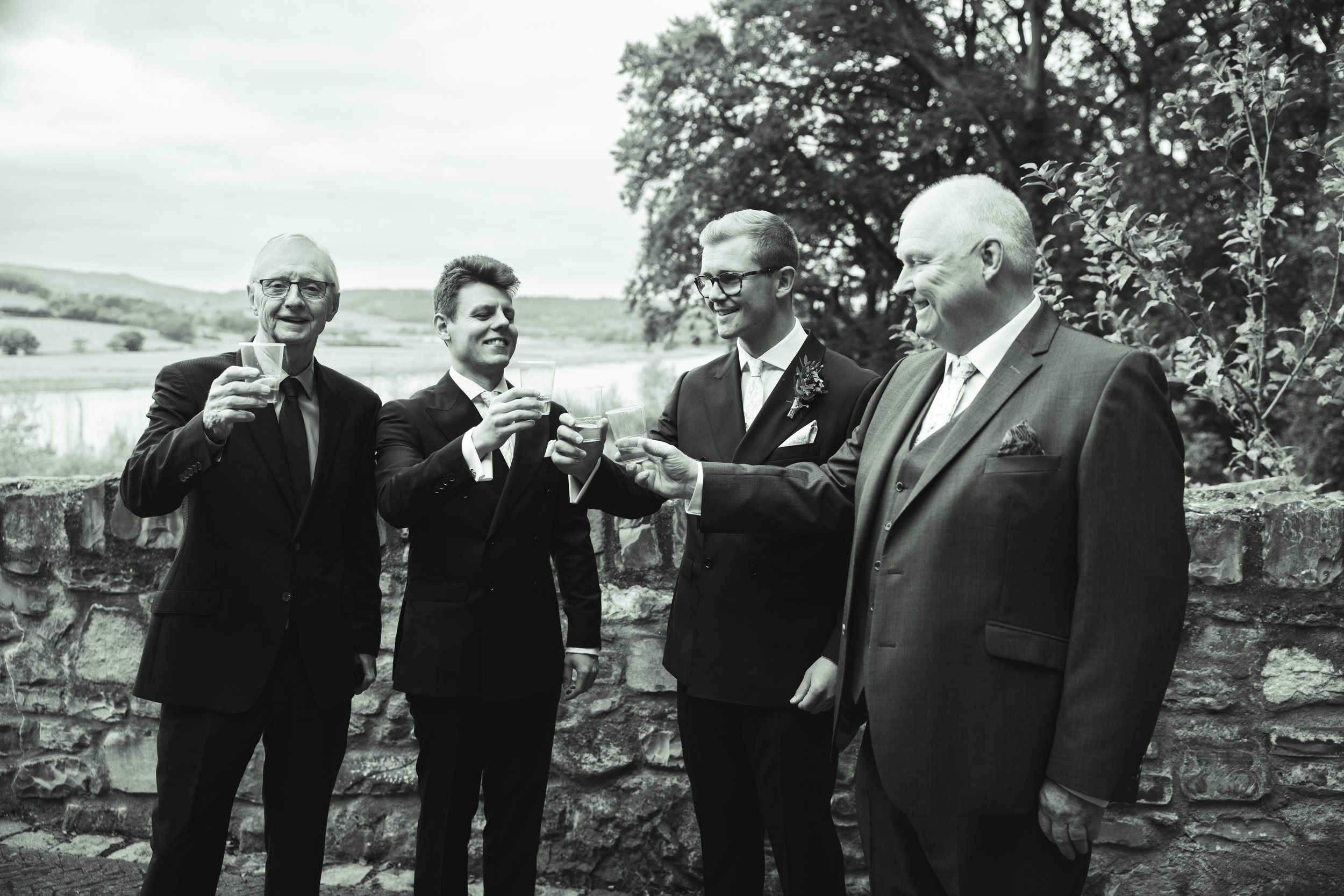 Five men in suits holding glasses, toasting outdoors near a stone wall at a wedding celebration.