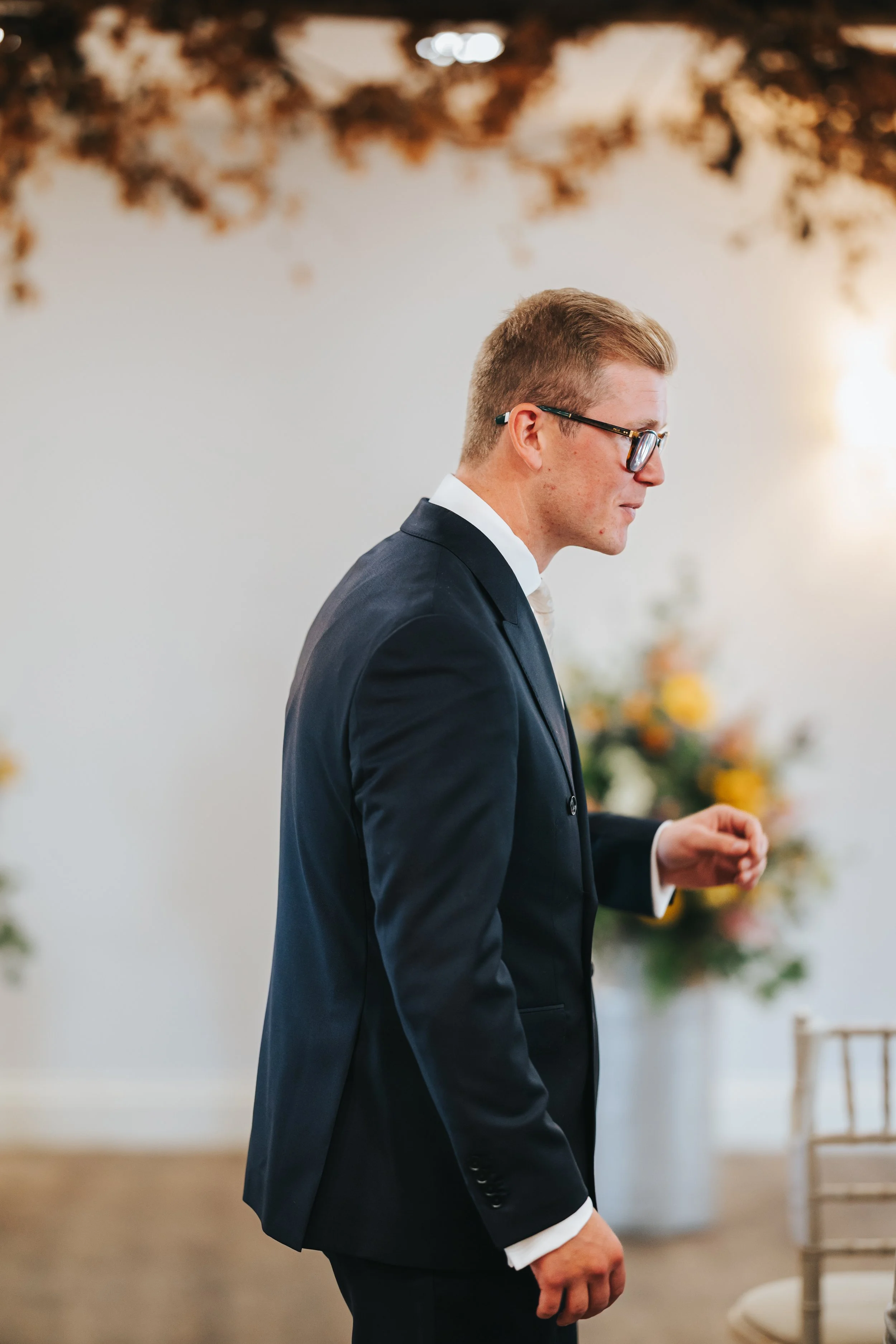A young man in a black suit and glasses standing indoors at a formal event, with floral arrangements in the background.