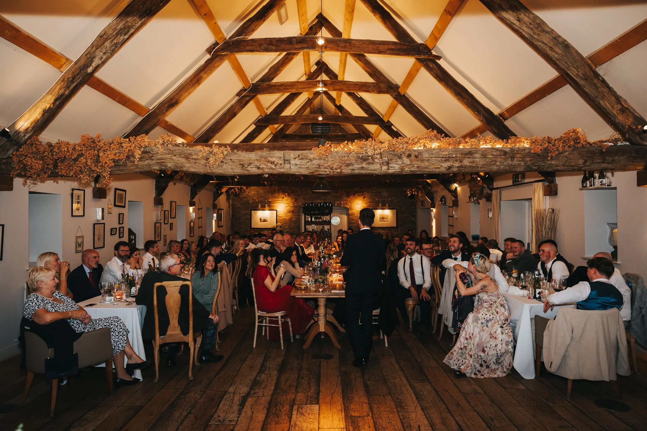 A wedding reception in a rustic venue with wooden beams, decorated with dried flowers, filled with guests seated at long tables, and a person in a black suit standing in the center.