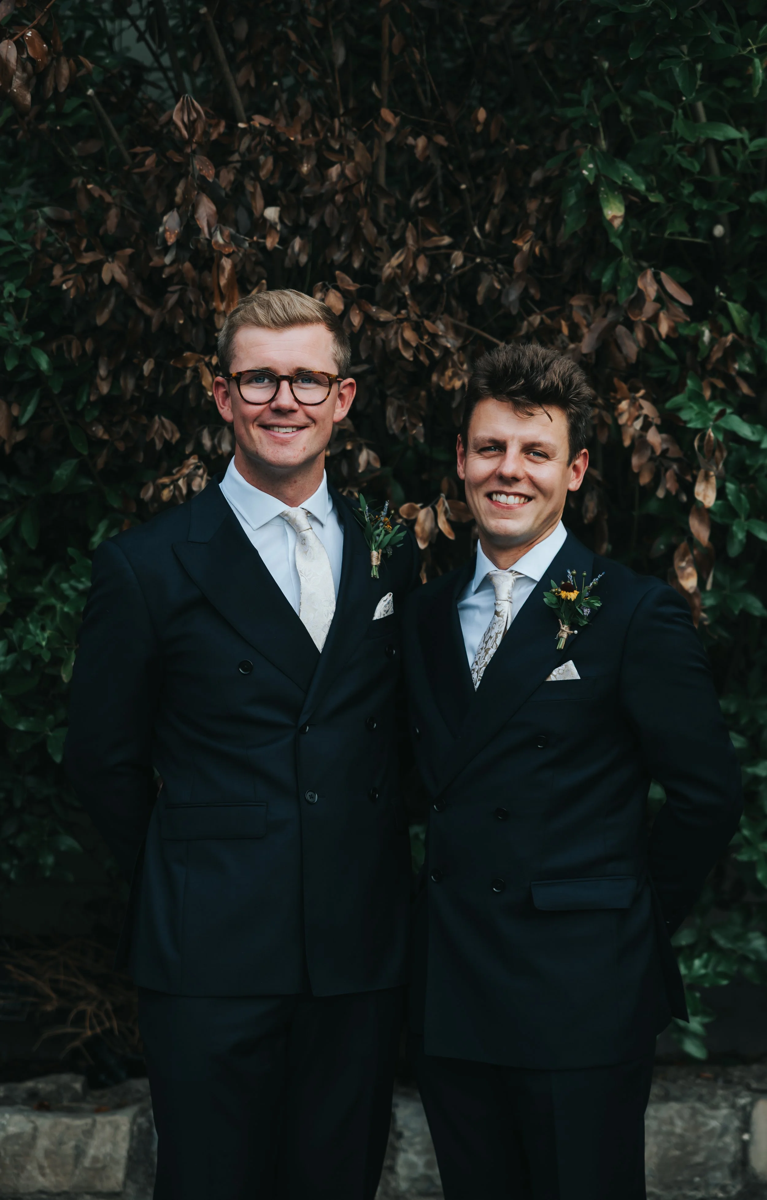 Two men wearing formal black suits with boutonnières, white shirts, and patterned ties, standing outdoors in front of a leafy background, smiling at the camera.
