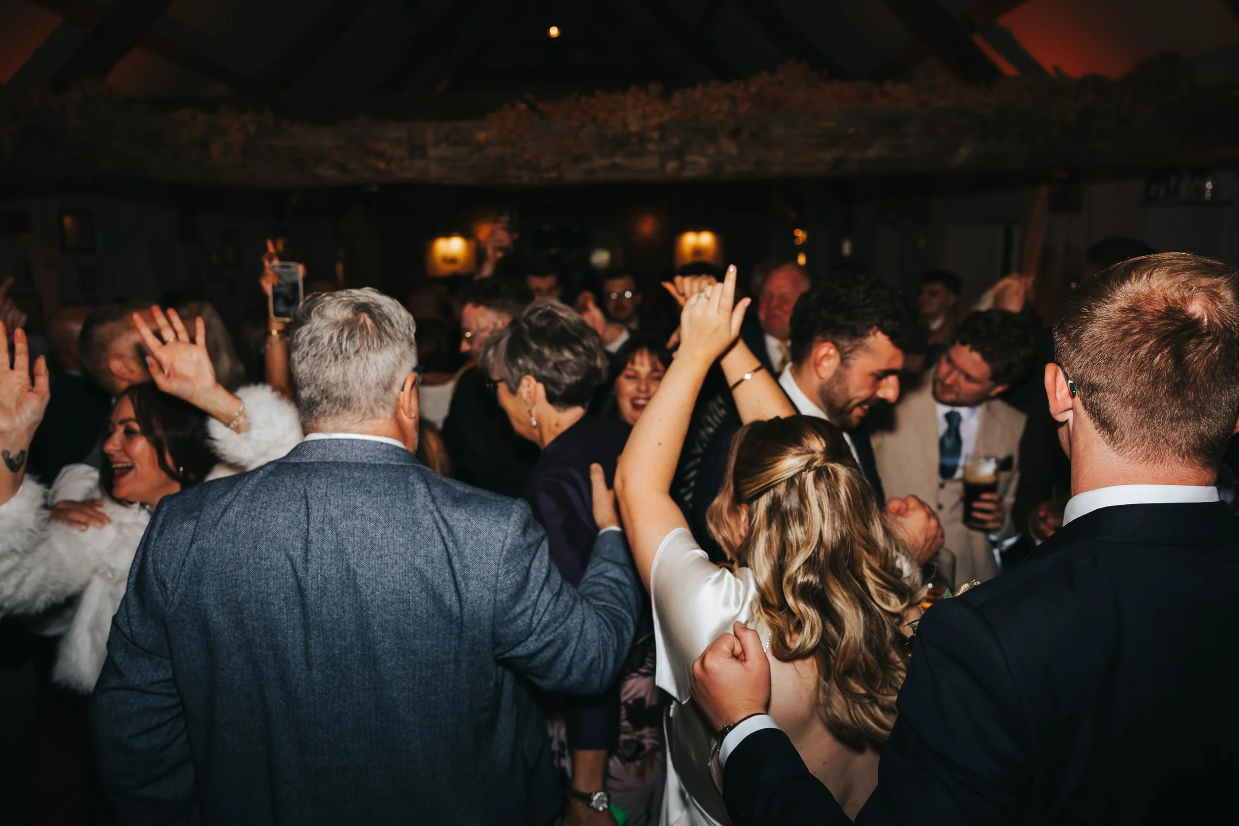 People dancing and celebrating at a lively party or wedding reception in a dimly lit rustic venue.