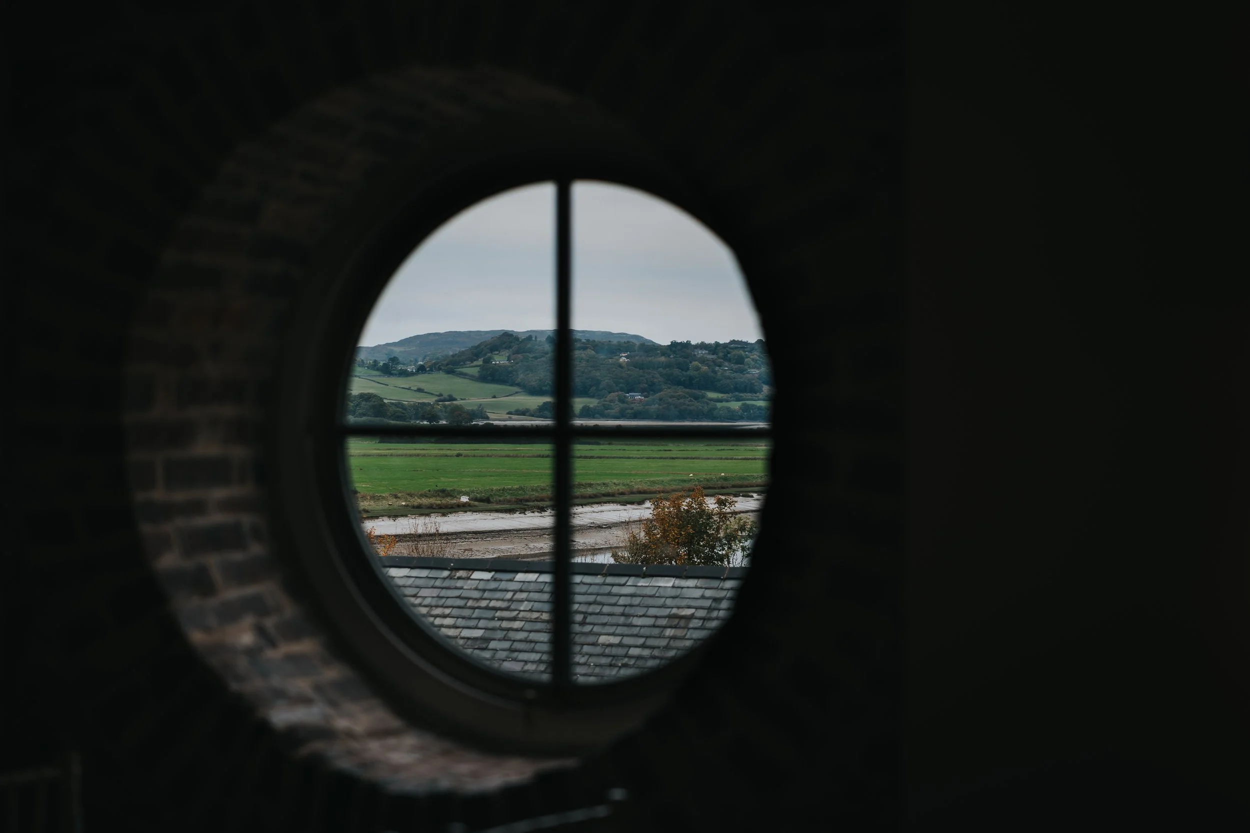 Landscape view through circular window with cross bars, showing green fields, a river, and hills in the distance.
