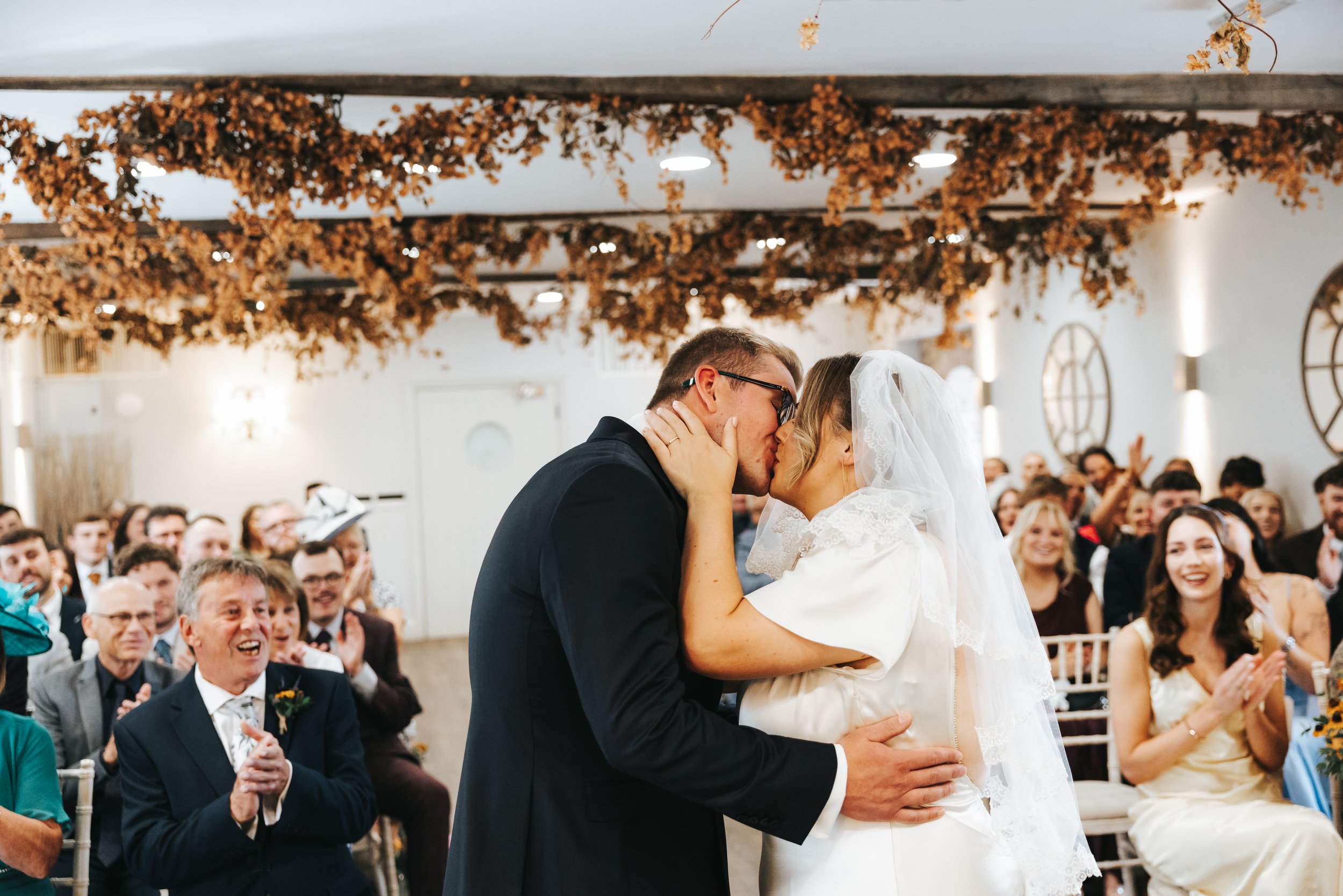 A newlywed couple sharing a kiss during their wedding ceremony in front of seated guests. The groom is wearing a black suit and glasses, and the bride is in a white wedding dress with a lace veil. The audience is smiling and clapping in the backgroun
