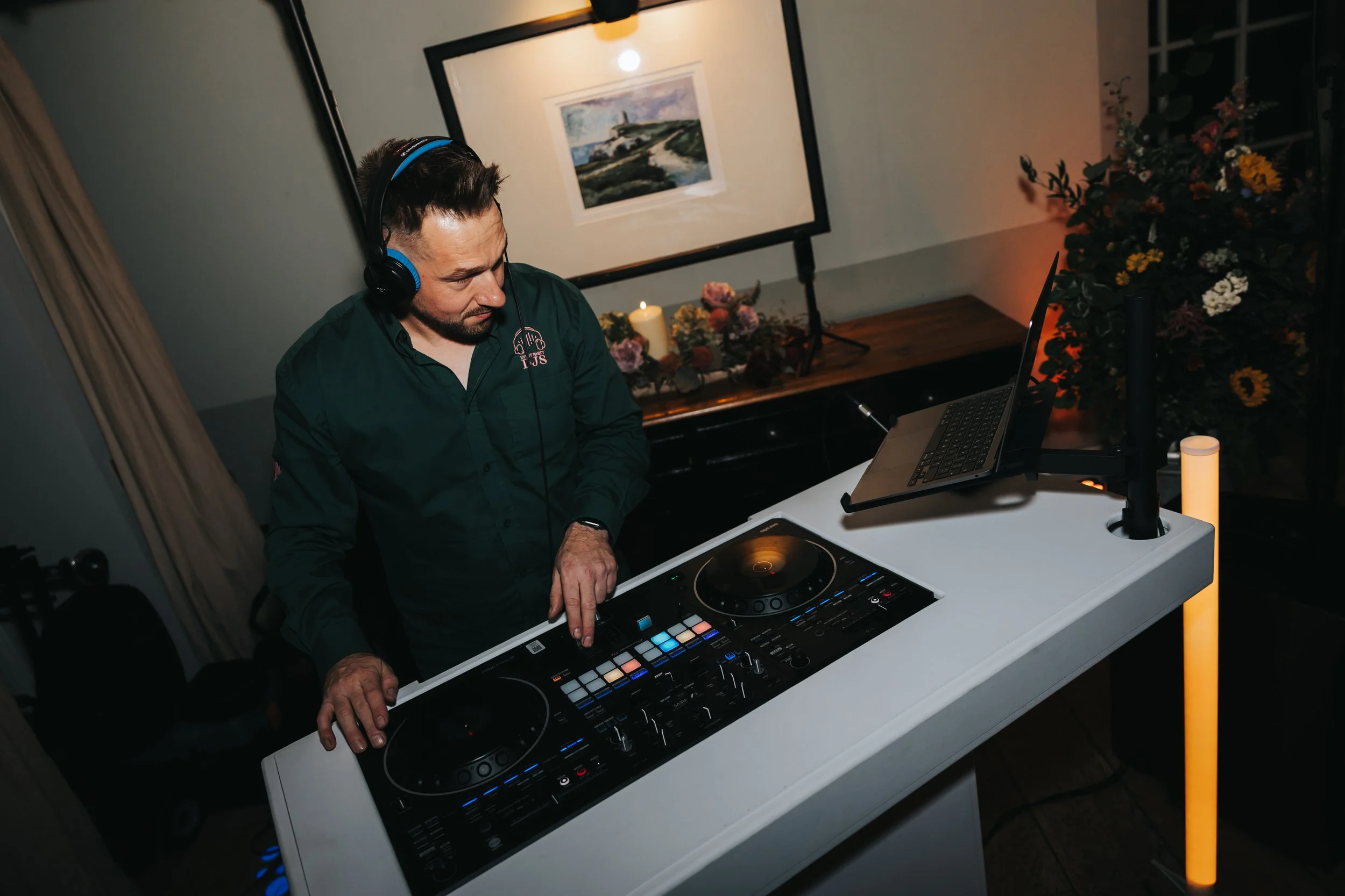A male DJ with headphones, wearing a dark green shirt, is operating a DJ controller and laptop at a white table, in a room with framed artwork, floral decorations, and a candle in the background.