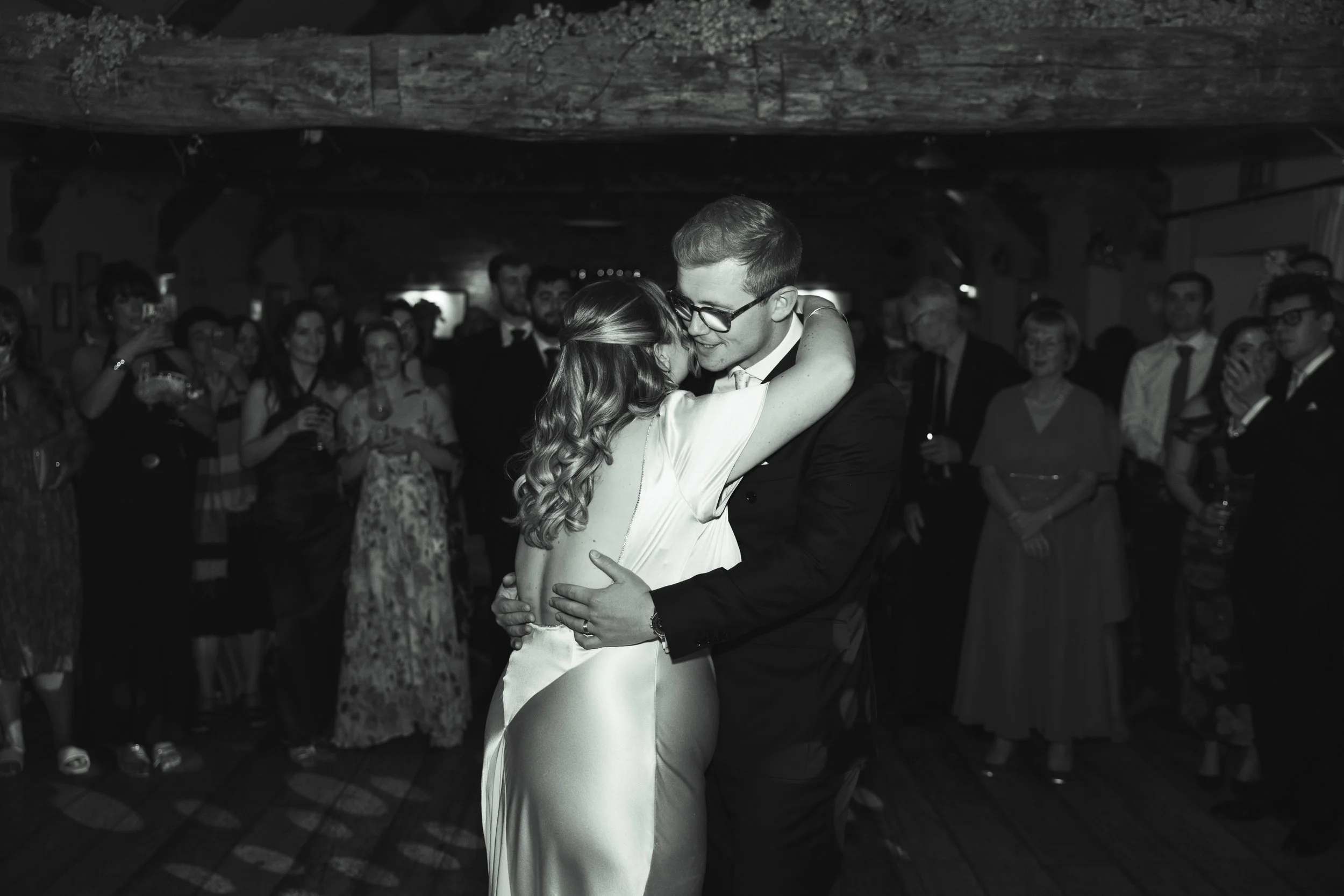 A couple is dancing closely at a wedding reception, surrounded by guests watching and taking pictures.