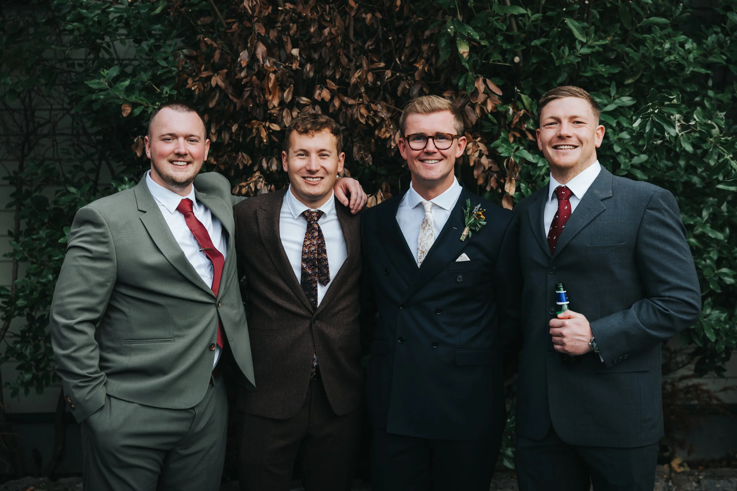 Four men in suits smiling and standing close together outdoors with lush green foliage in the background. One man is holding a bottle.