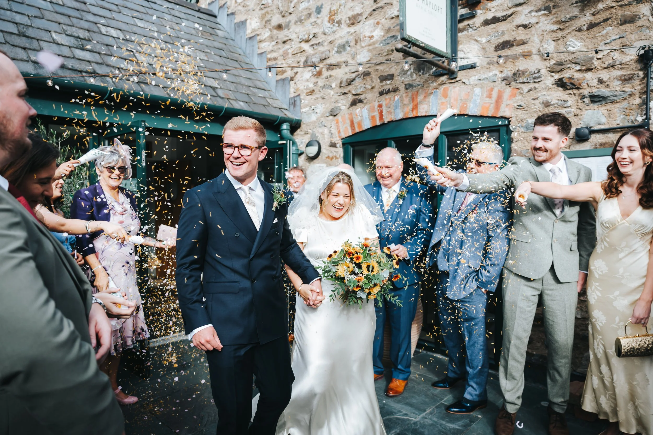 A joyful bride and groom holding hands and smiling, surrounded by friends and family in wedding attire, celebrating with confetti outside a rustic stone building.