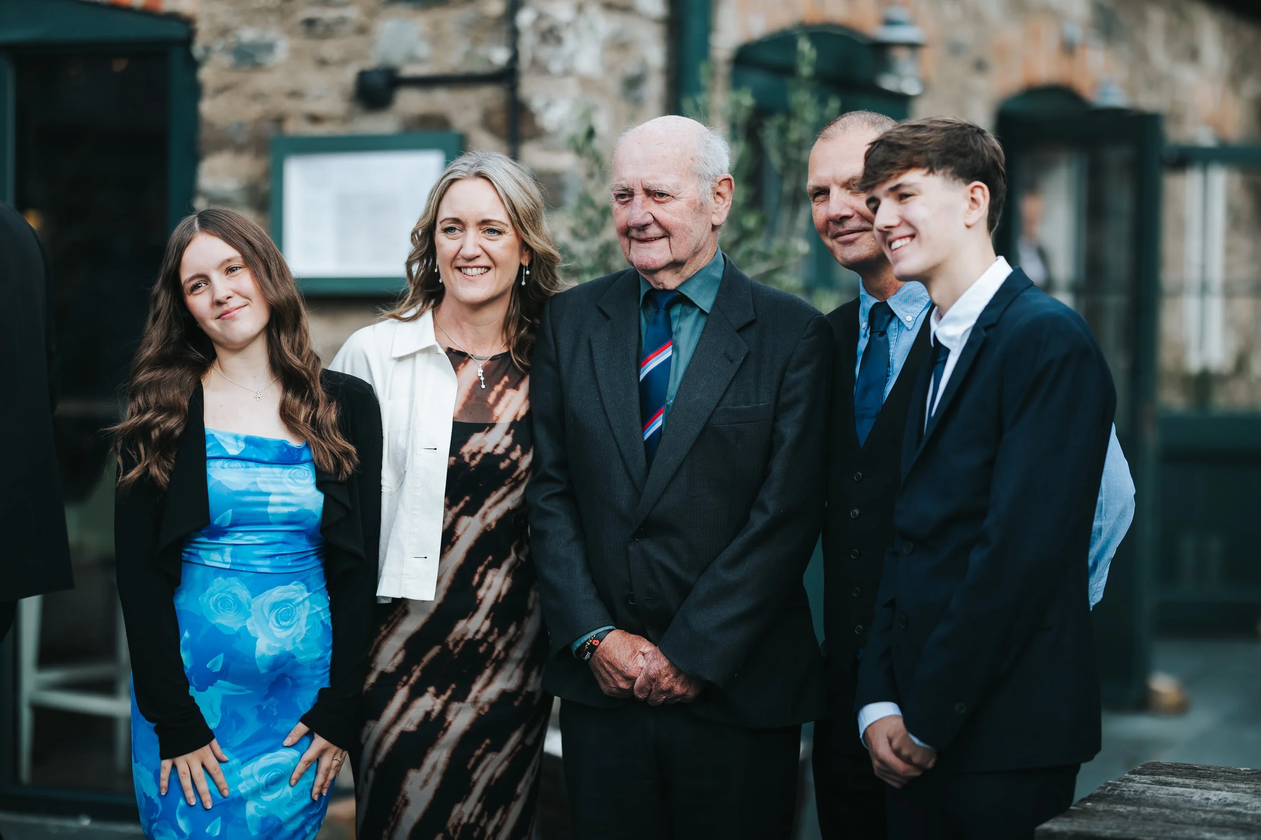 Group of five people standing together outdoors, smiling and looking in the same direction, in front of a brick building.