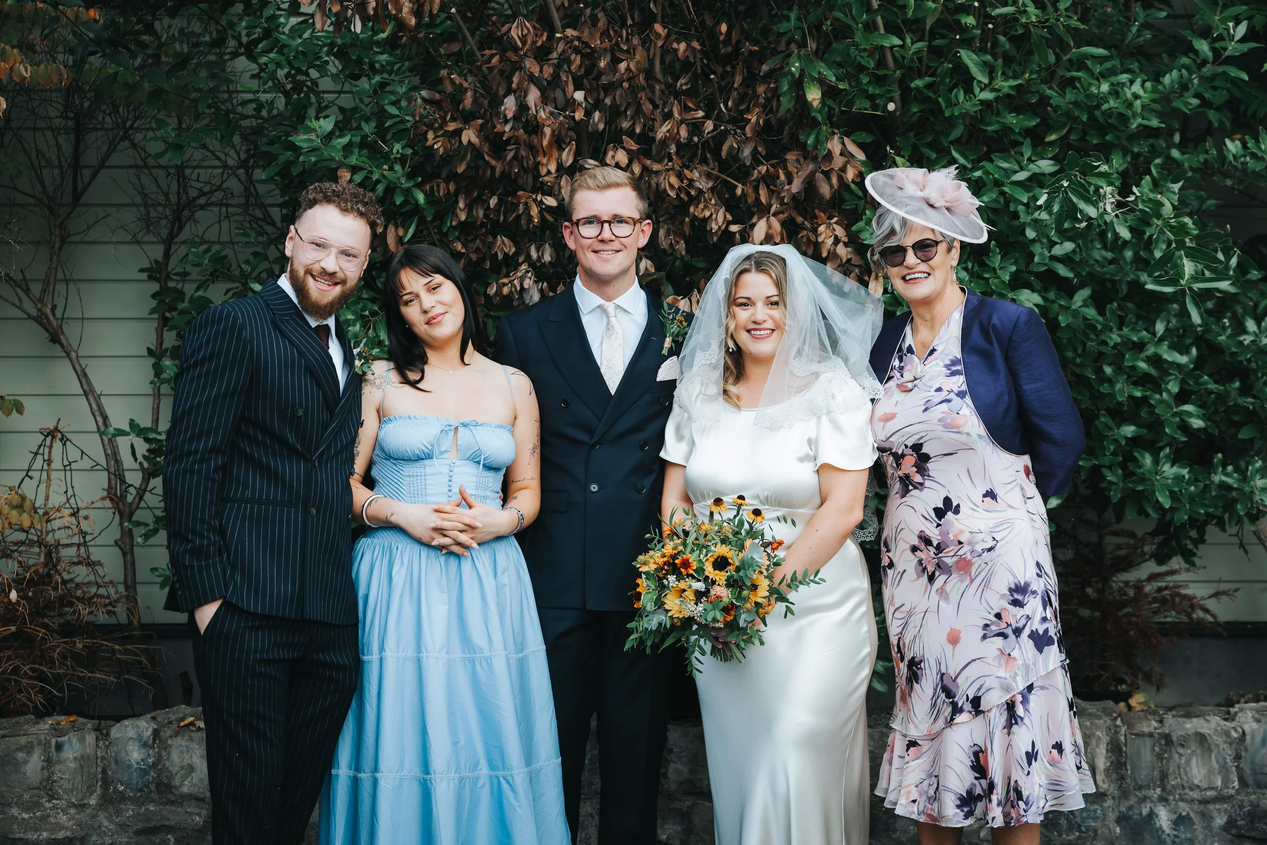 Group of six people smiling at a wedding, including bride in white dress with veil holding a bouquet of flowers, groom in navy blue suit, and four other adults in formal attire standing outdoors in front of greenery.