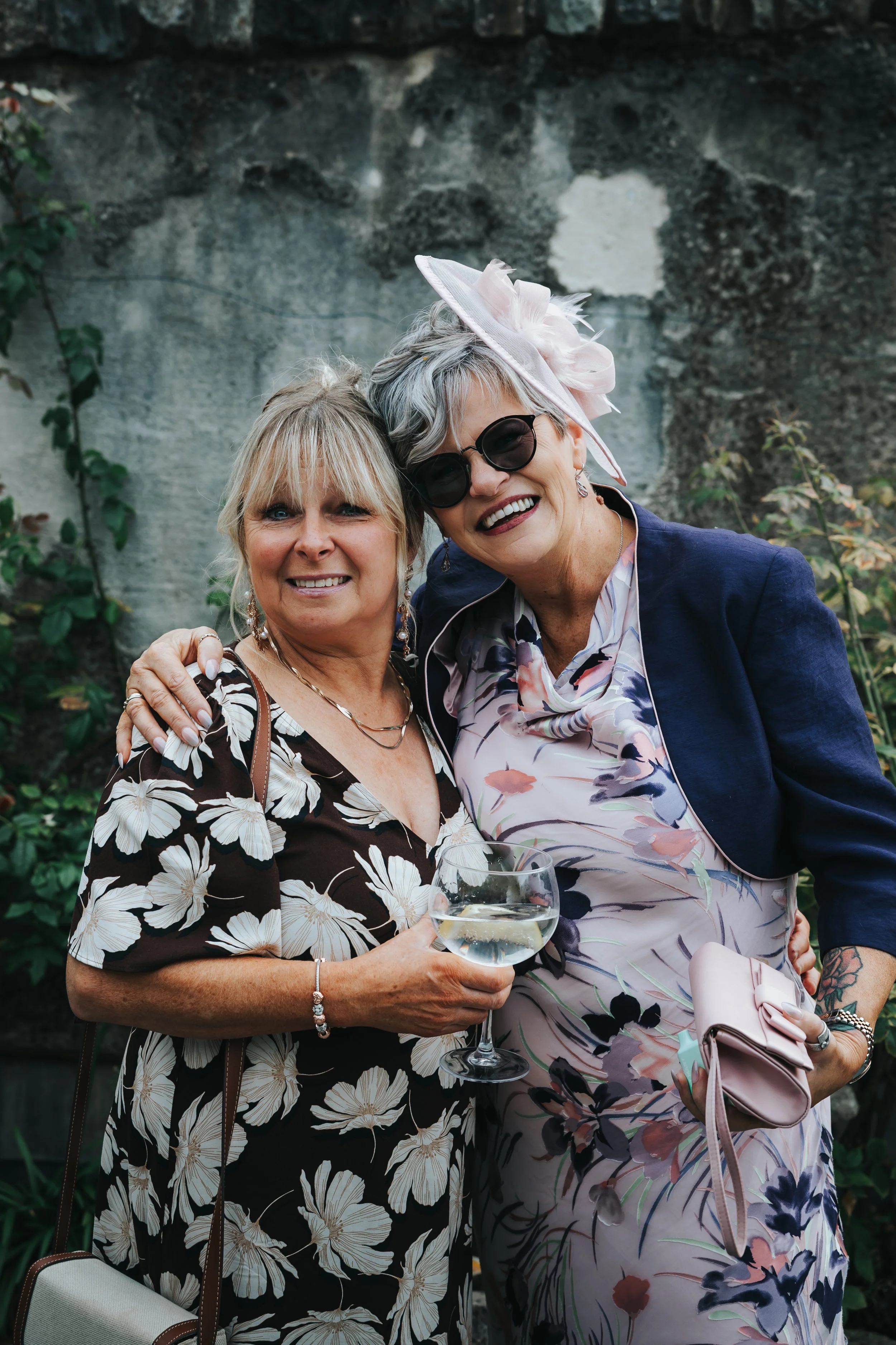 Two women smiling at an outdoor event, one holding a glass of wine, the other has a pink purse, both dressed in floral attire, with one wearing a large pink hat and sunglasses.
