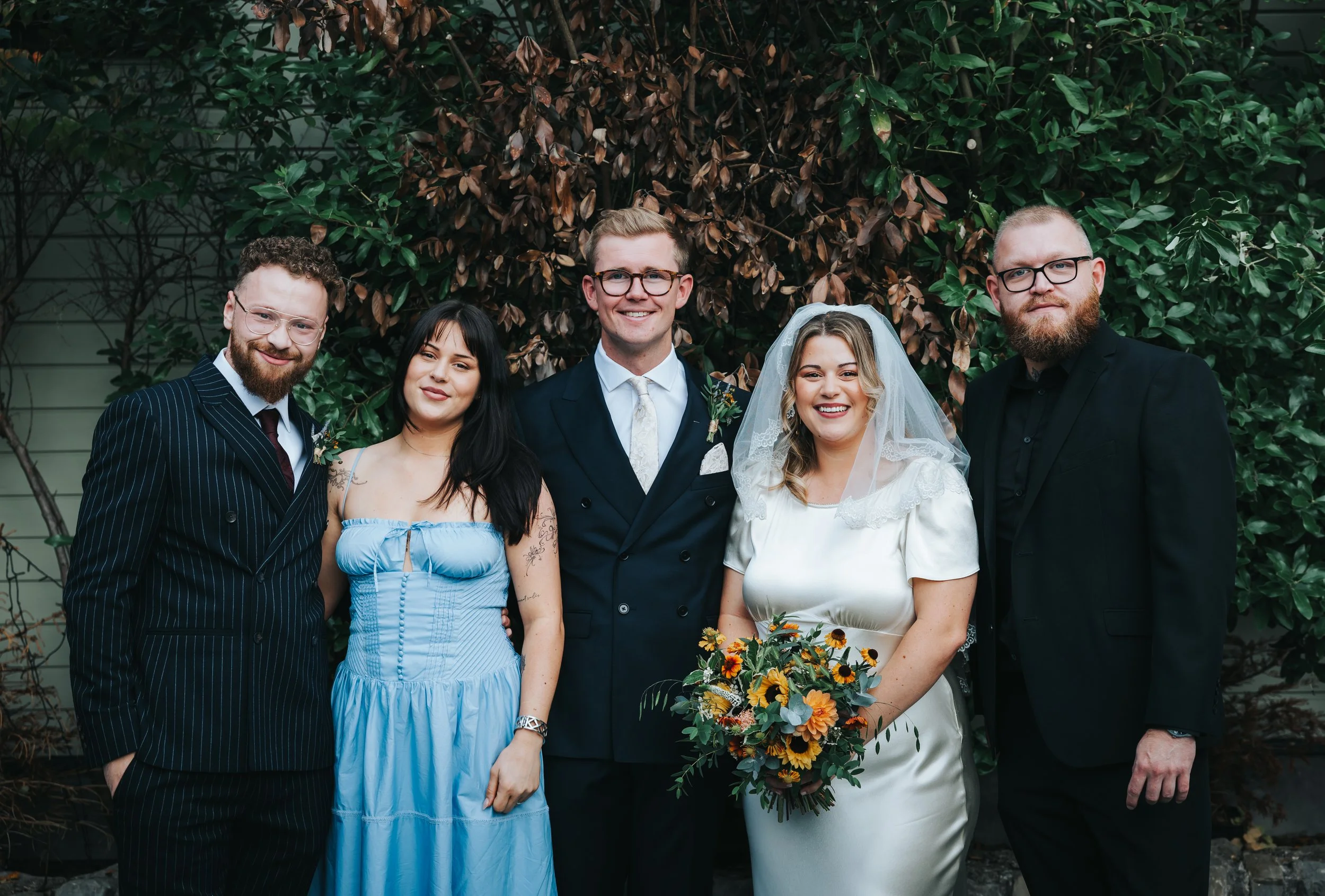 Group of six people, including bride and groom, standing outdoors in front of green and brown foliage. The bride is holding a bouquet of flowers and wearing a white satin dress and veil. The groom is wearing a dark suit and tie. The other four people