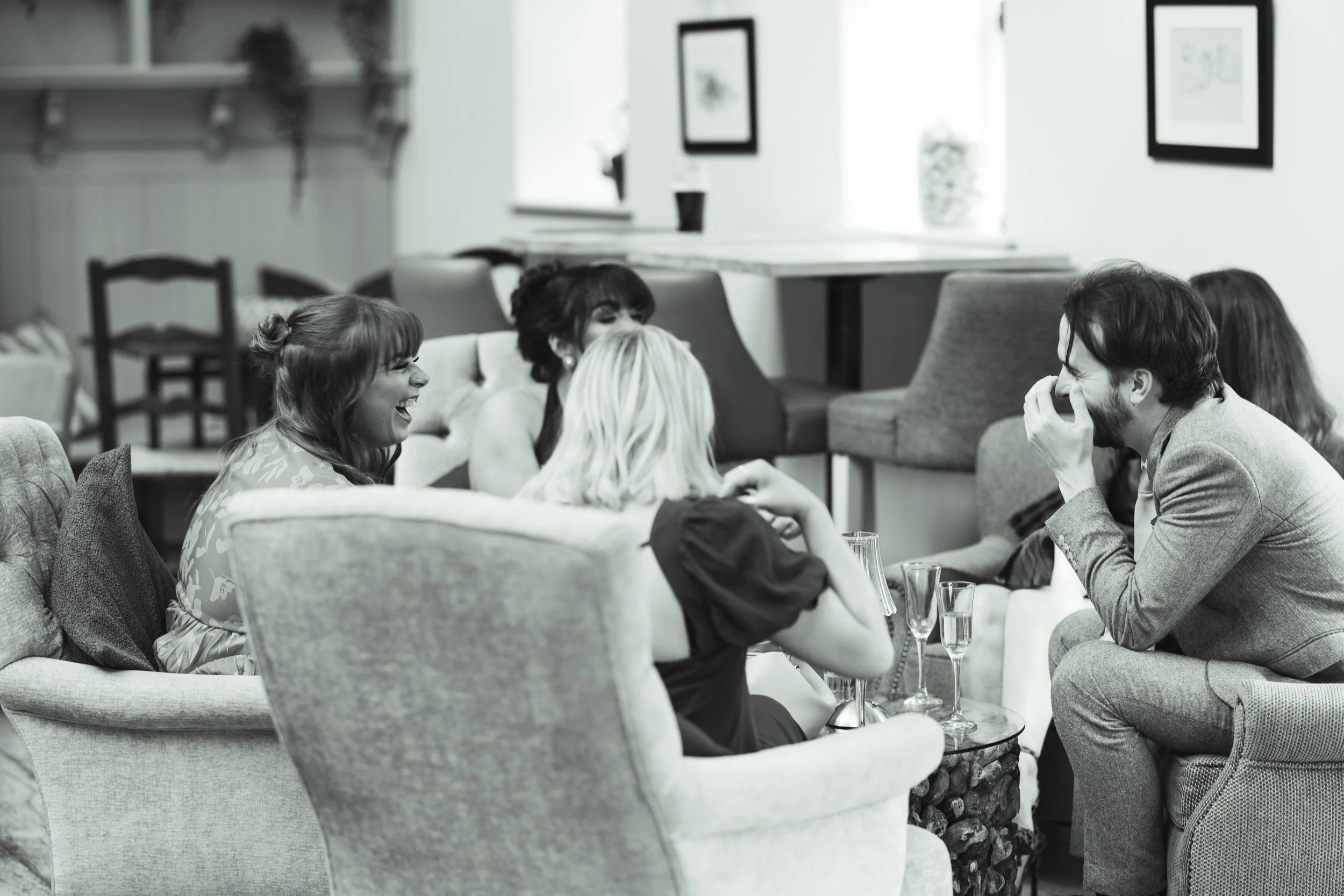 Group of friends laughing and enjoying drinks while sitting on a sofa and armchairs in a cozy living room.