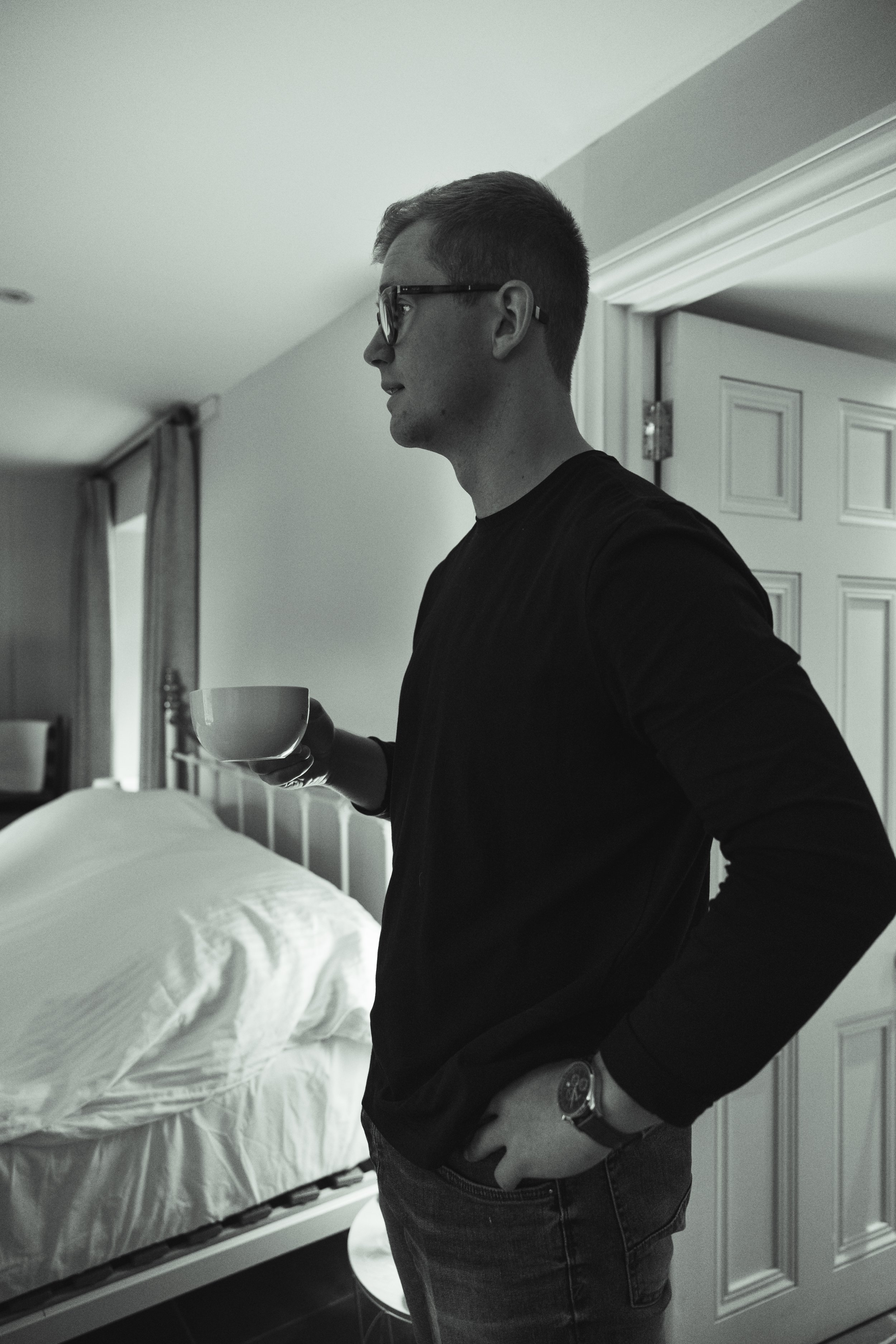 A man with glasses holding a cup, standing in a bedroom near a bed with white bed linens, in a black and white photo.