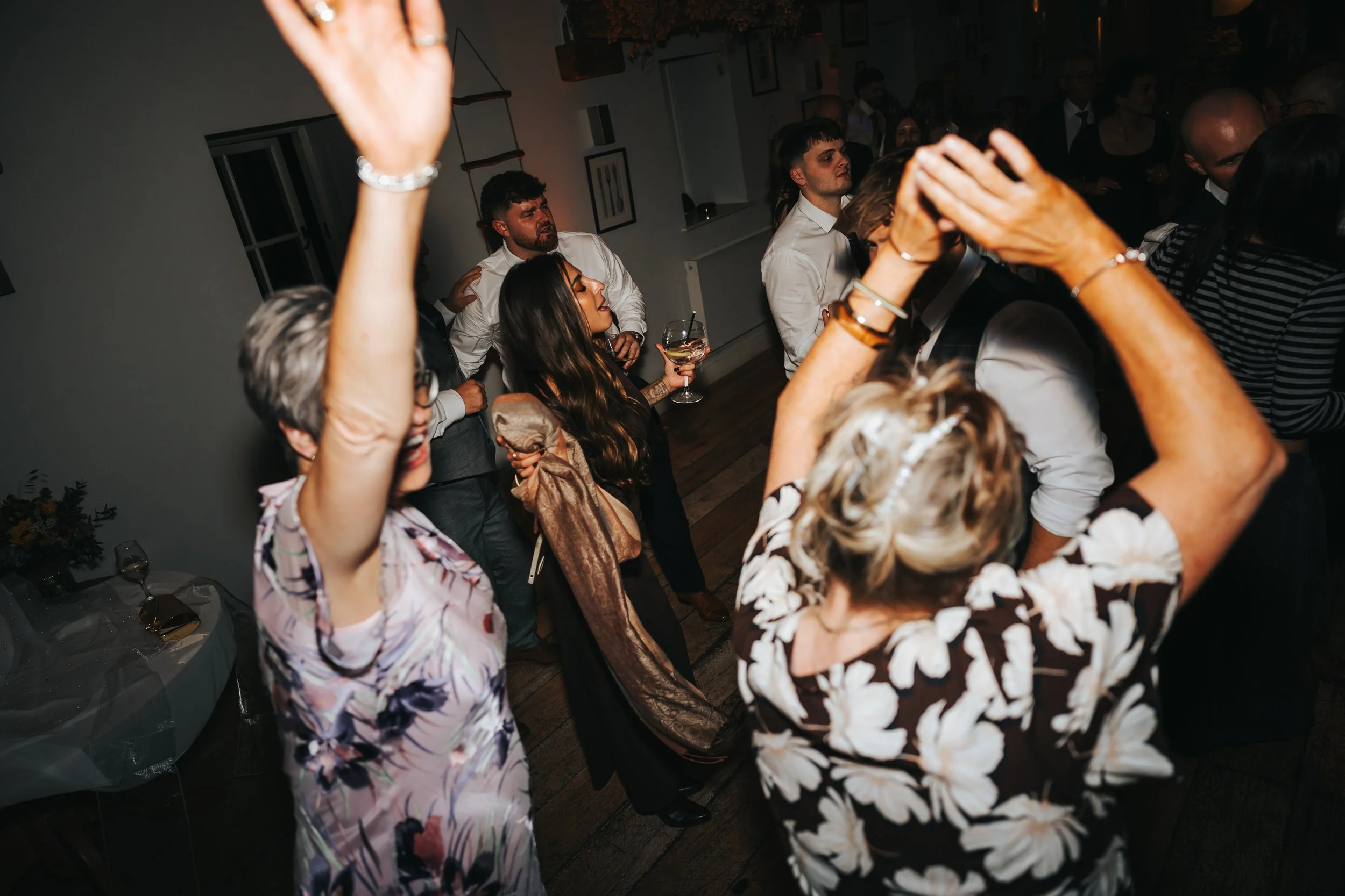 People dancing at a celebration or party indoors, with women and men in formal attire, some holding drinks, enjoying the music and dancing.