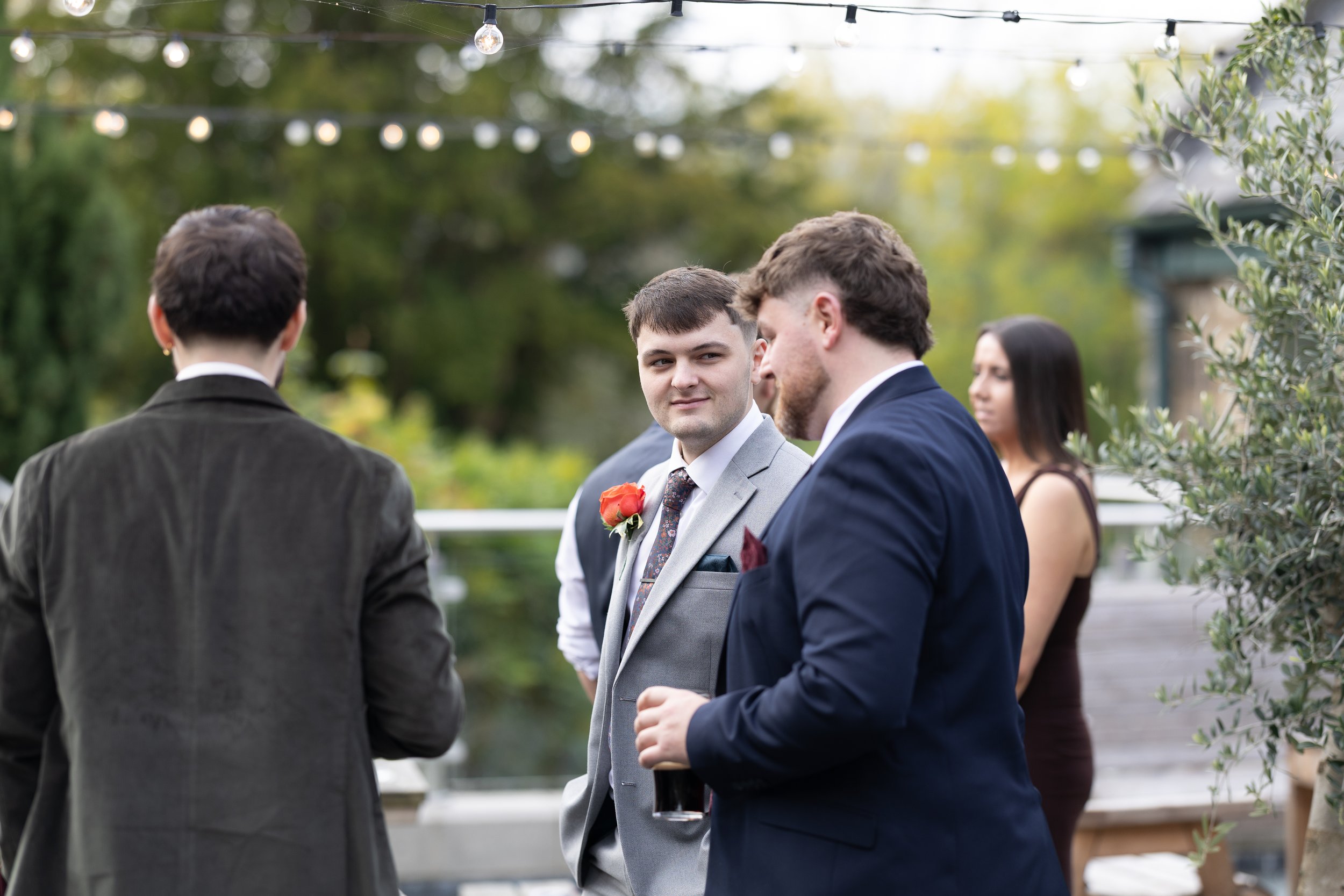 Group of people in formal attire chatting outdoors at an event with string lights overhead and greenery in the background.