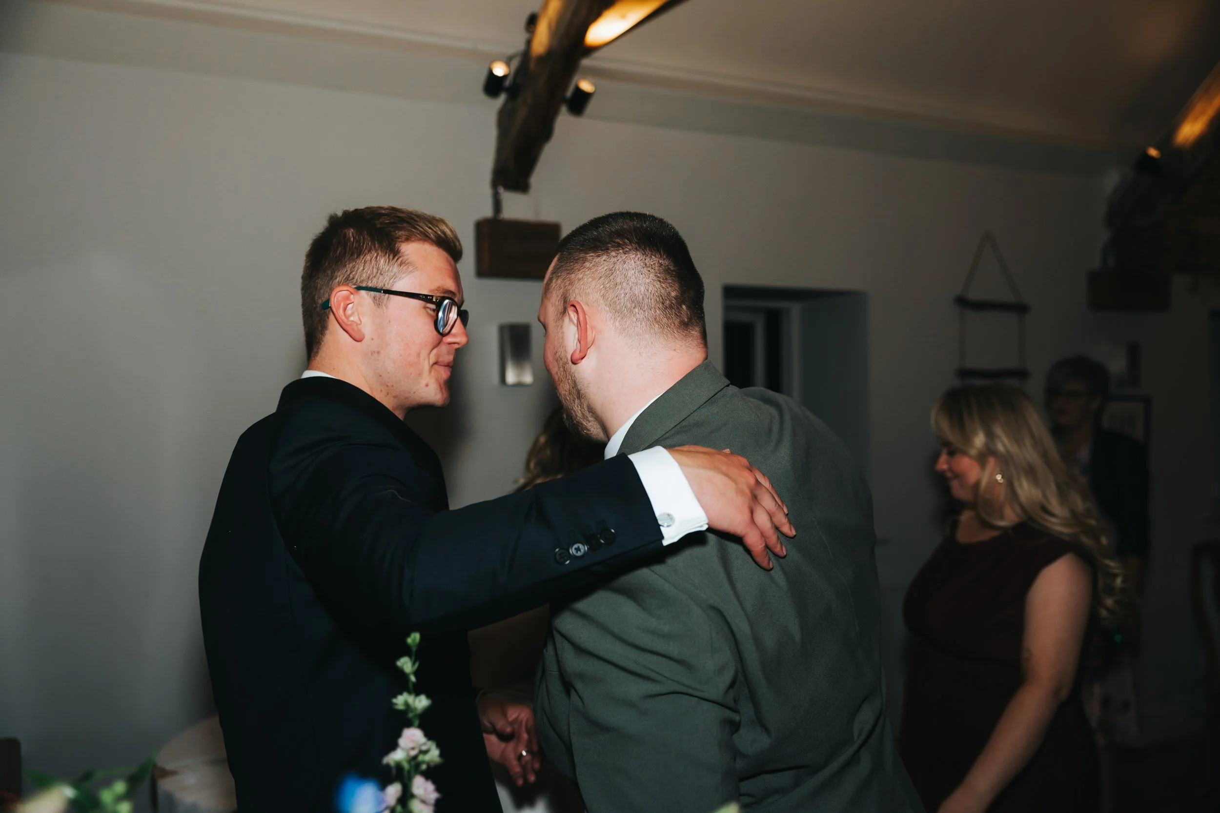 Two men in suits embrace and smile at each other during a social gathering, with women in the background.