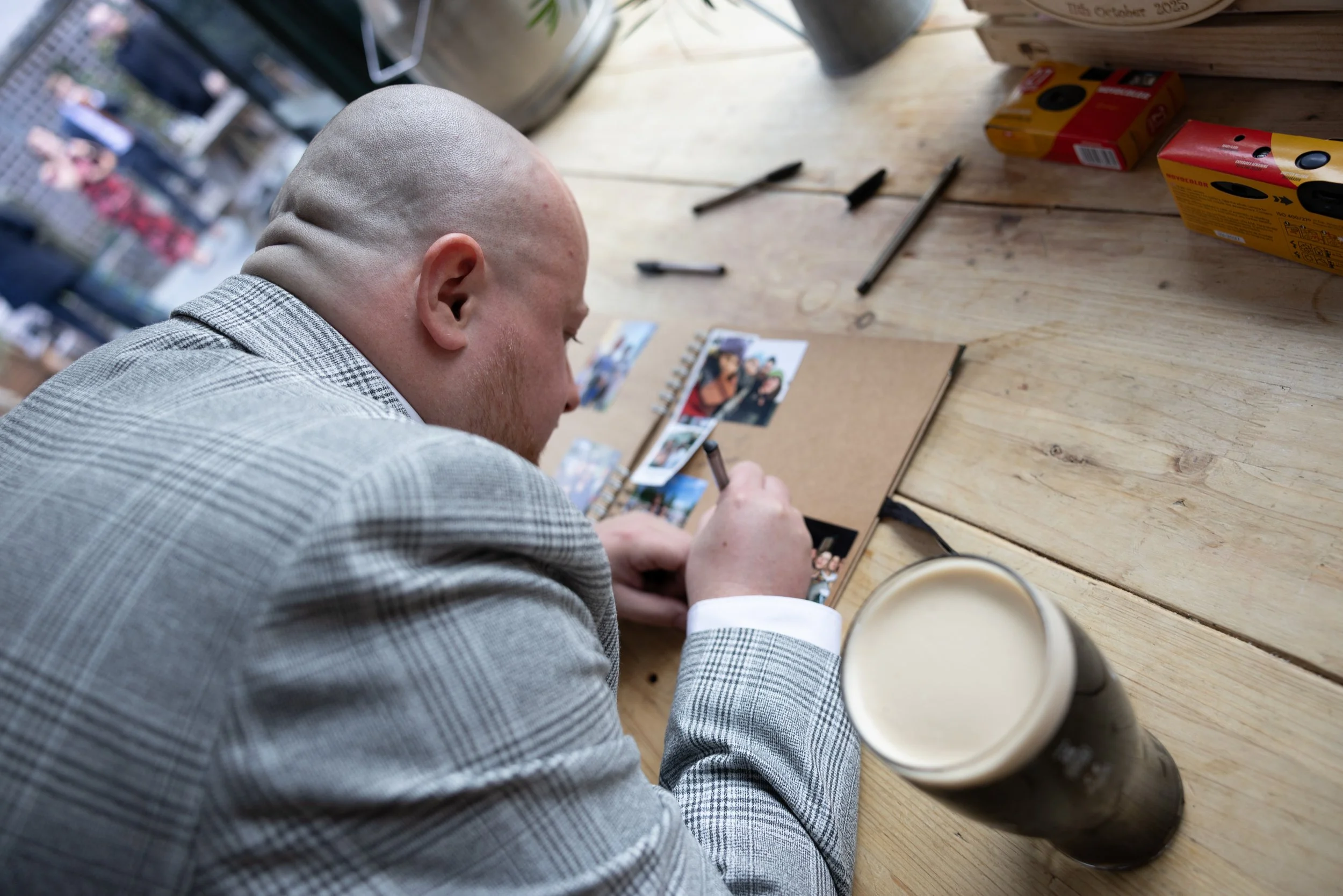 A bald man wearing a gray plaid suit is sitting at a wooden table, signing a photo album surrounded by pens, with a pint of dark beer in the foreground.