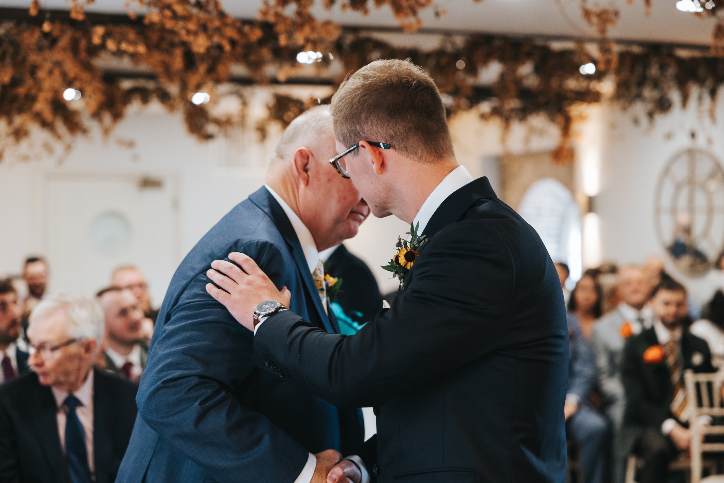 A wedding ceremony where a young man in a black suit with a sunflower boutonniere is touching foreheads with an older man in a blue suit, surrounded by seated guests in a decorated venue.