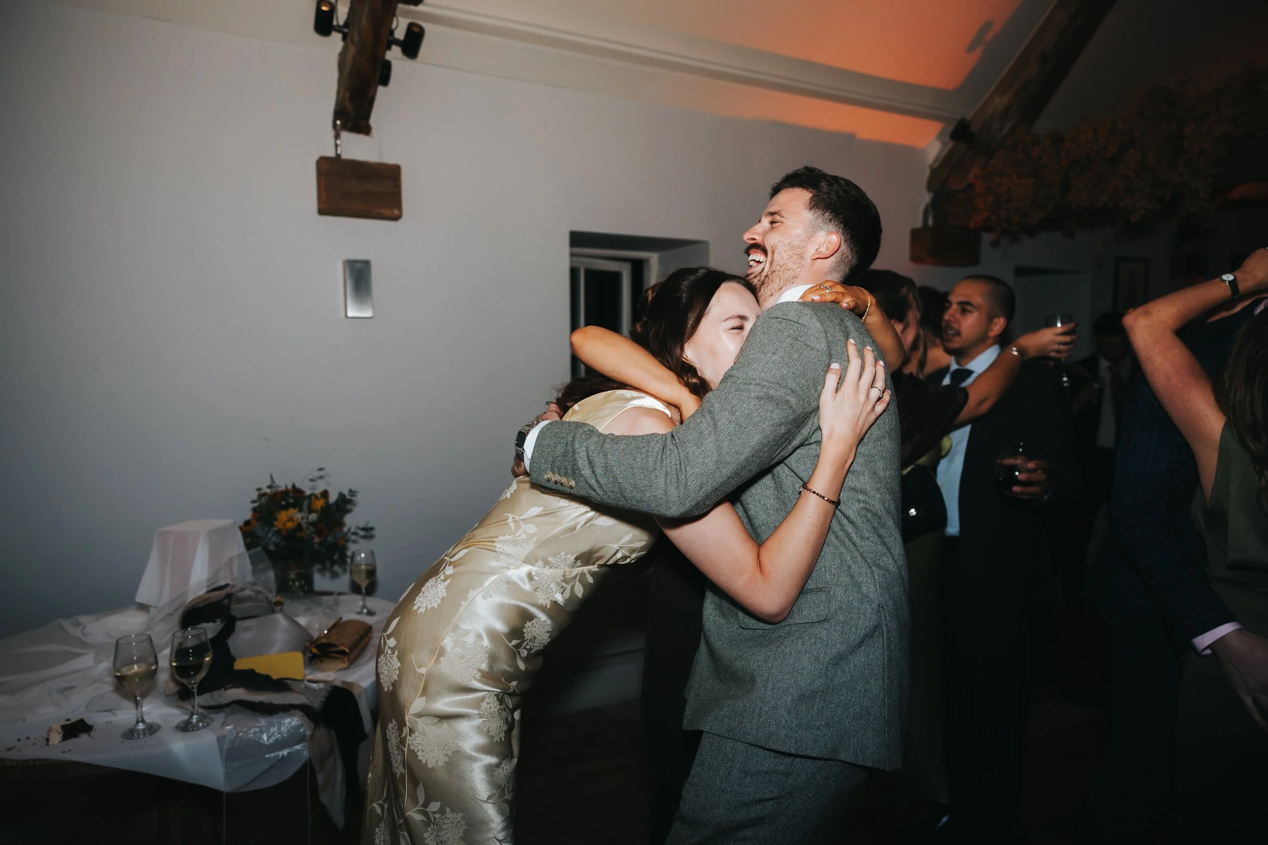 A couple sharing a joyful hug at a celebration, with other guests in the background holding drinks.