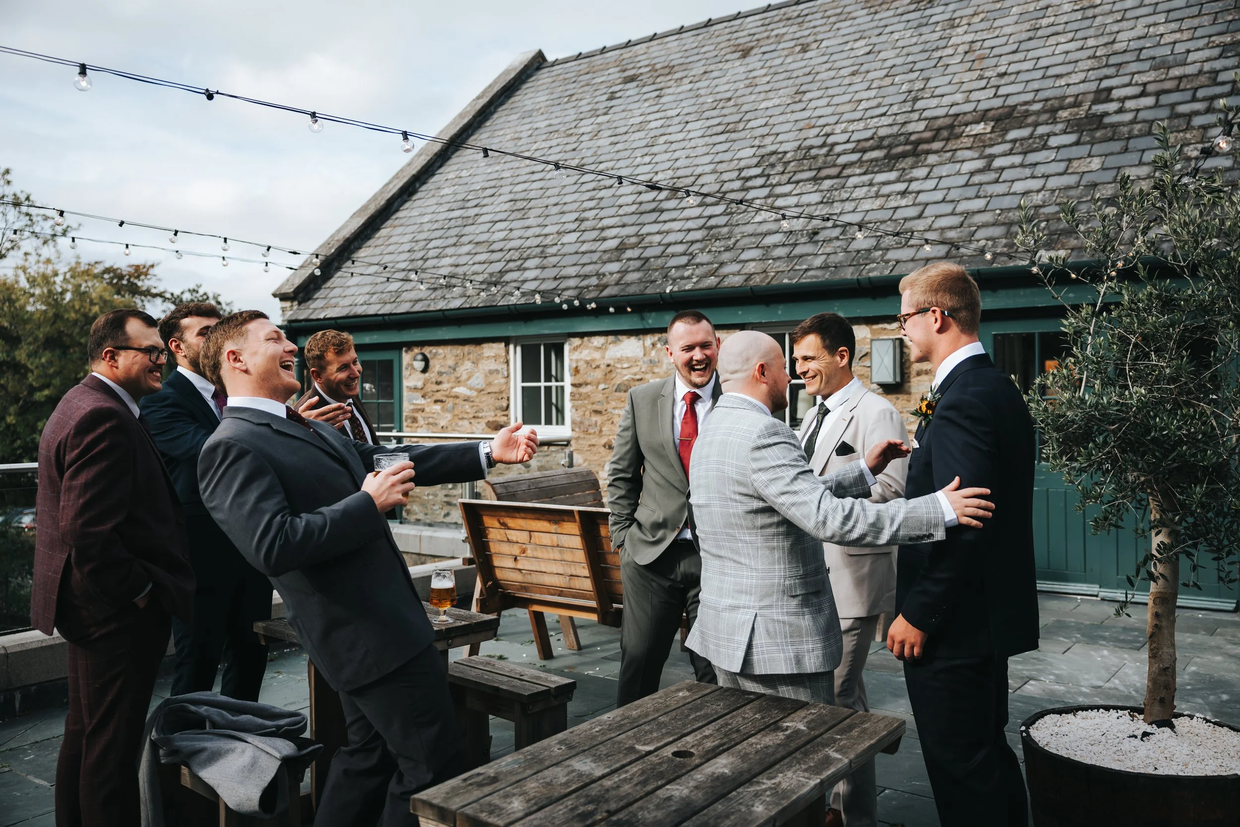 Group of men in suits socializing outdoors on a rooftop patio with string lights, a stone building, and potted plants.