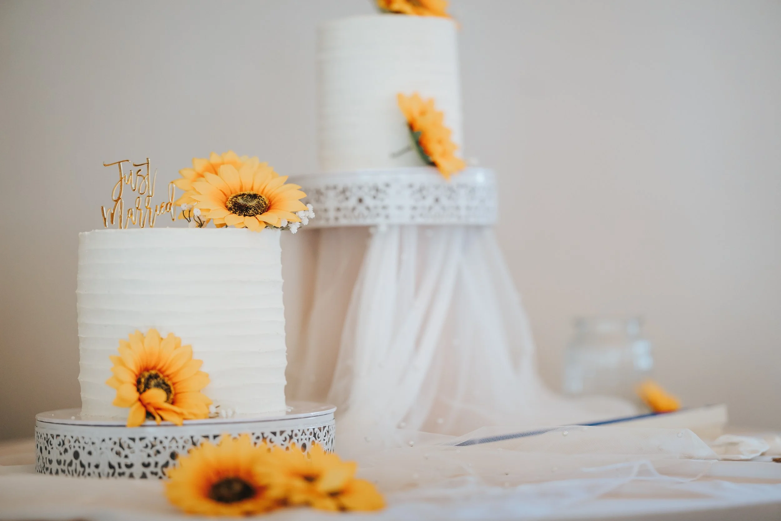 Wedding cake decorated with sunflower flowers, with a 'Just Married' topper, on a table with a white cloth, and a blurred cake in the background.