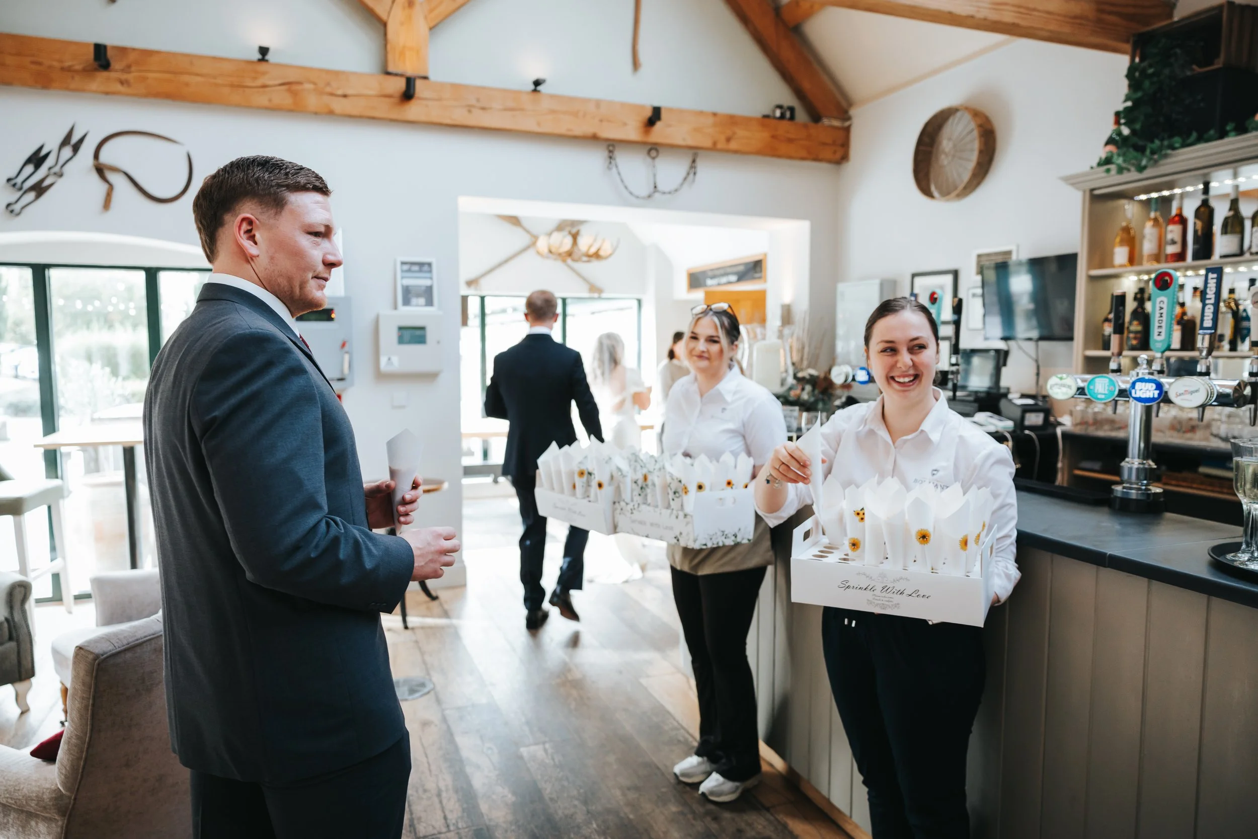A man in a suit at a bar or restaurant interacting with two smiling women holding boxes of small gift bags or favors, with other patrons in the background.