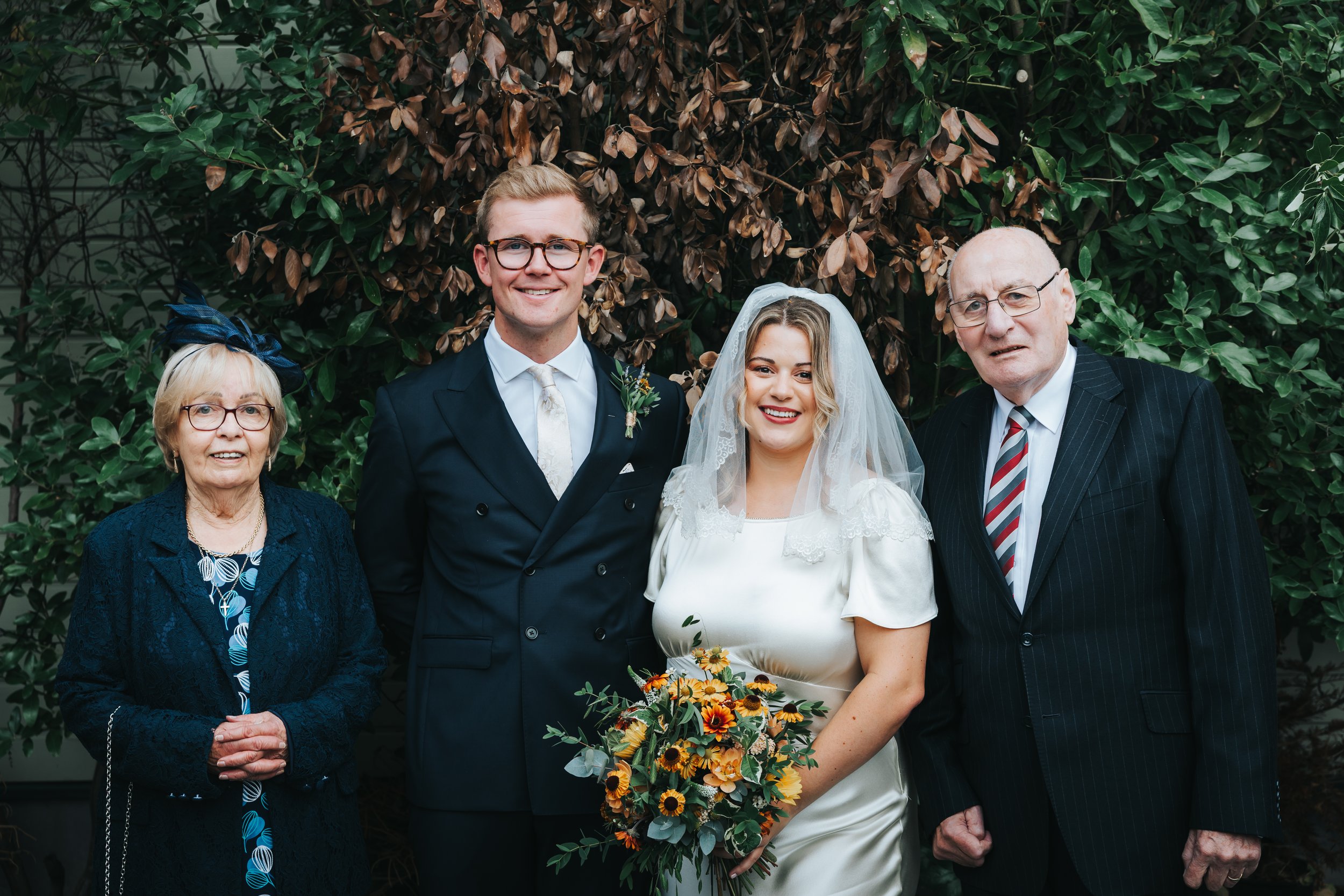 A group of five people at a wedding, standing outdoors in front of green foliage. The bride is in a white dress with a veil, holding a bouquet of flowers. The groom is in a dark suit with a light tie. An elderly woman in a dark blue outfit and hat st
