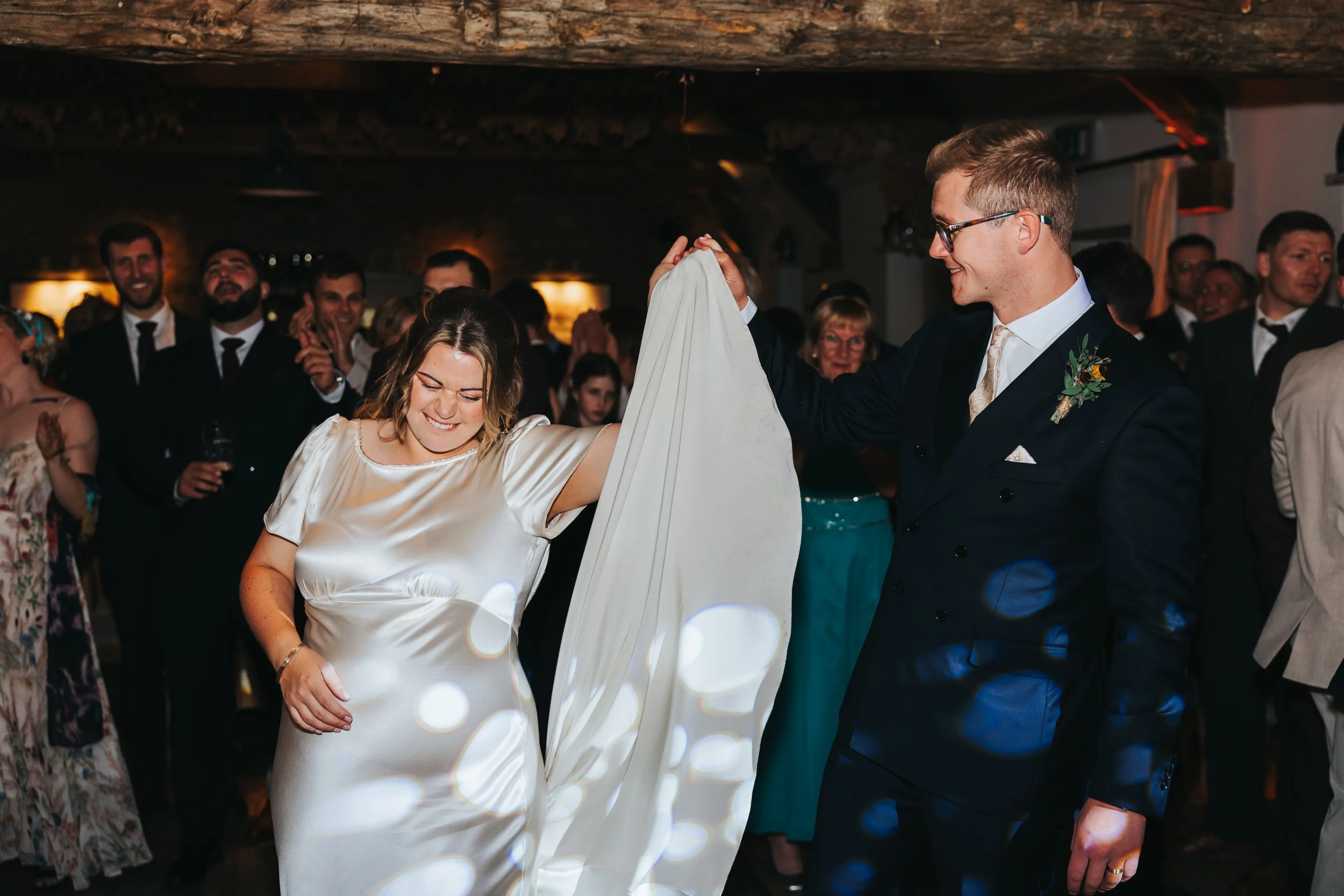 A bride and groom dancing at a wedding reception. The bride is wearing a satin wedding dress and smiling, while the groom is in a dark suit and holding her hand. Guests are clapping and watching in the background.