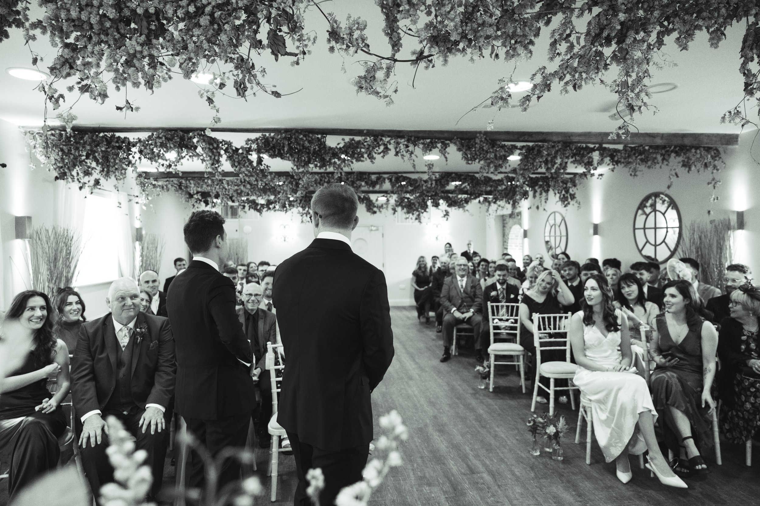 Two men in tuxedos give a speech at a wedding reception. Guests sit on chairs, smiling and listening. The room is decorated with flowers and circular wall art.