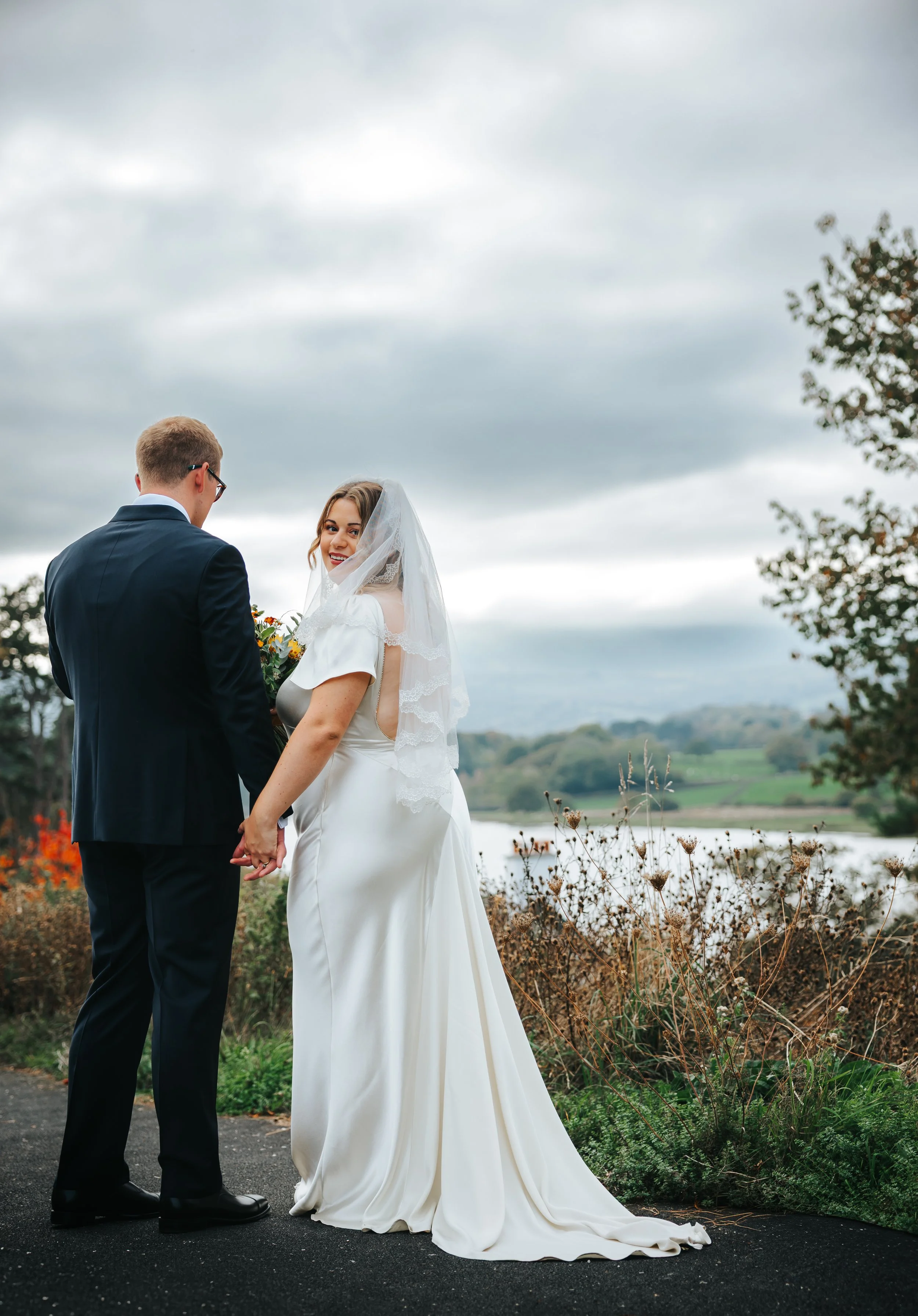 A bride and groom standing outdoors near a river, holding hands, with the bride smiling at the camera and wearing a white wedding dress and veil, and the groom wearing a dark suit, overcast sky in the background.