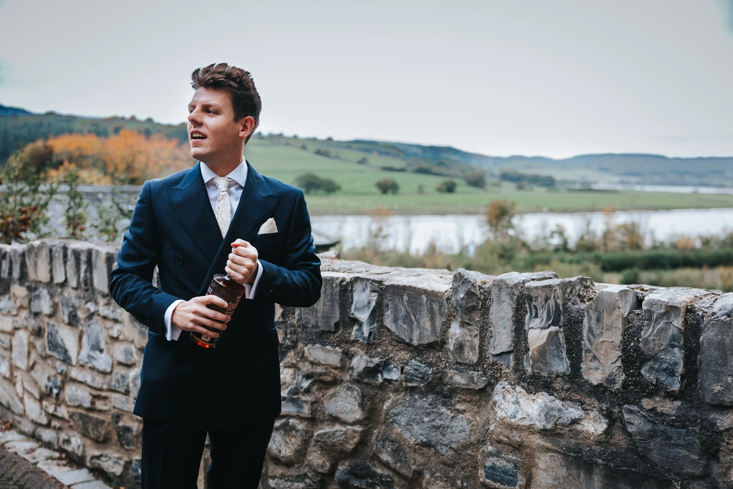 Young man in a formal navy suit and tie standing outdoors near a stone wall with a scenic view of a river and rolling hills in the background.