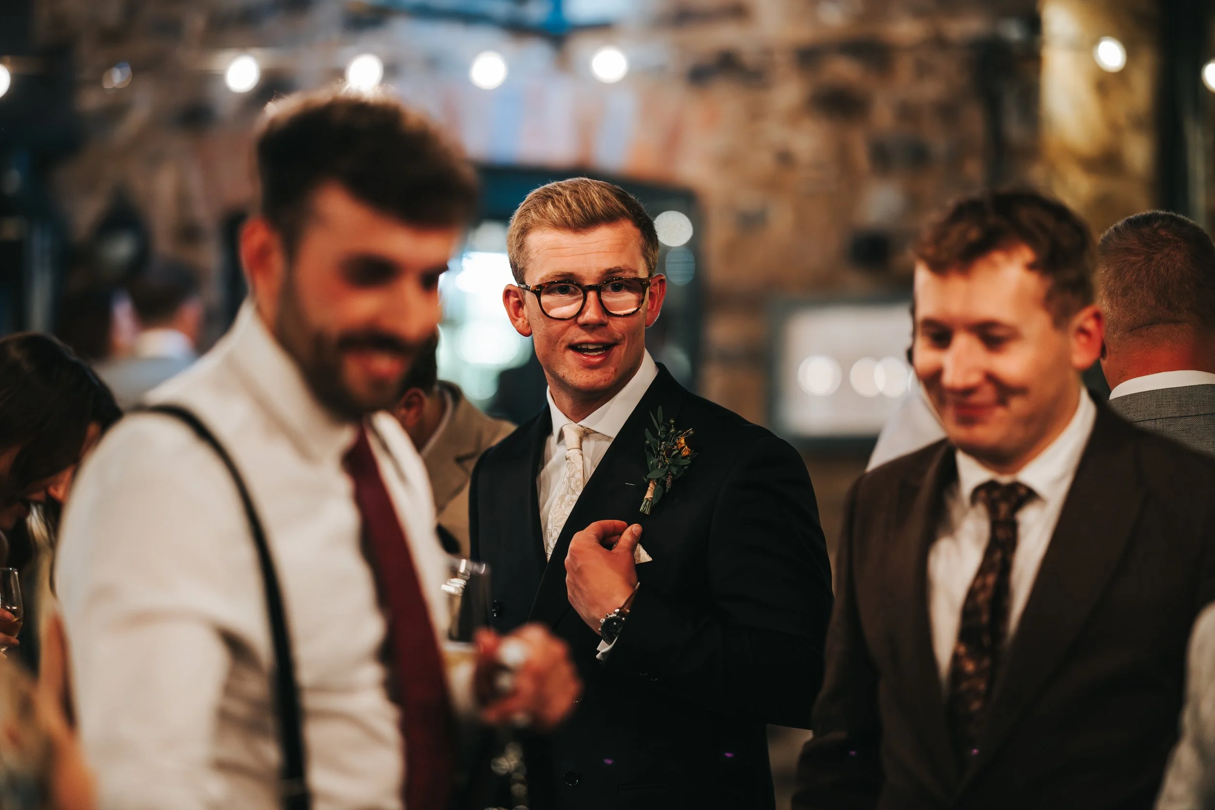 A group of well-dressed men at an indoor social event, with one man in the center wearing a suit, glasses, and a boutonniere, smiling and looking at someone off camera.