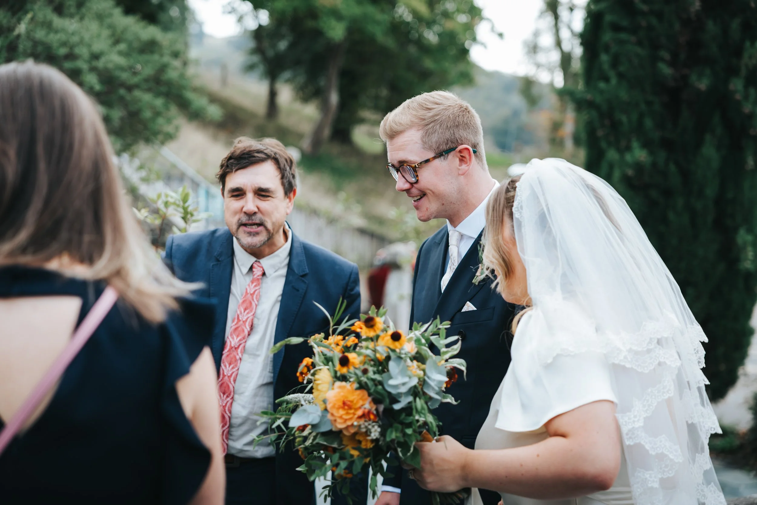 A bride and groom with a woman, possibly an officiant, standing outdoors on a wedding day, the bride holding a bouquet of flowers, with trees and a grassy hillside in the background.