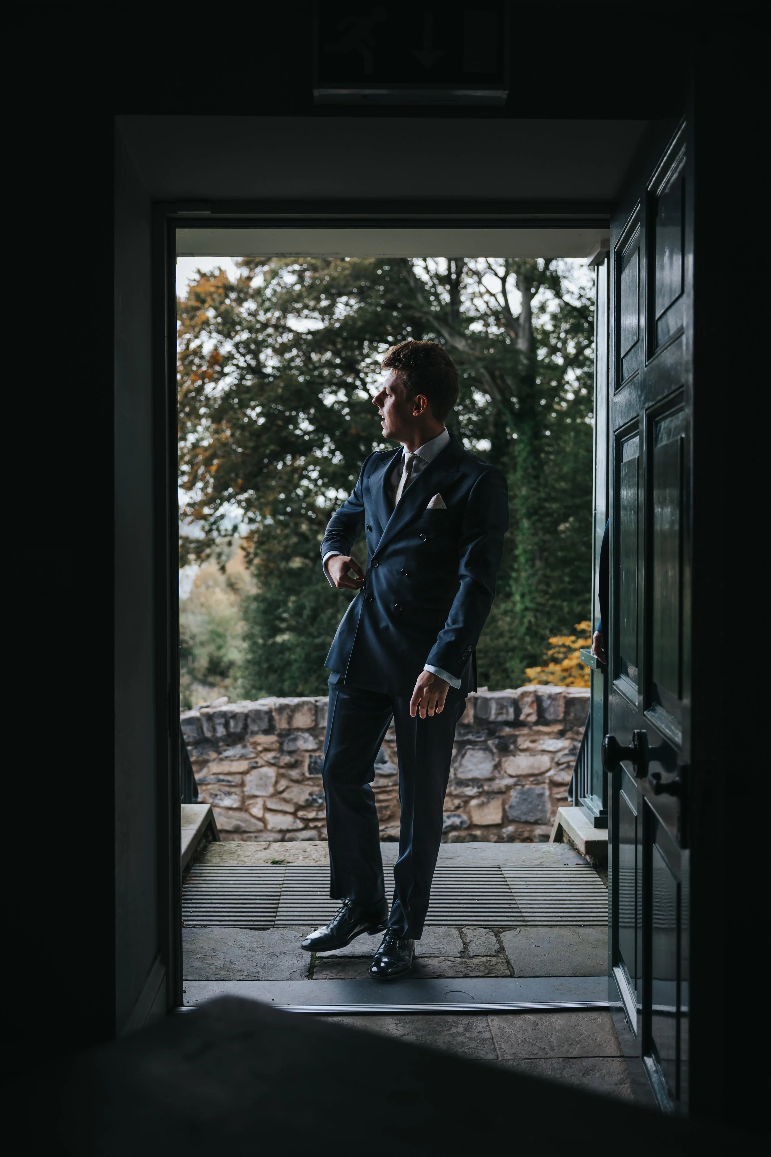 A man in a dark blue double-breasted suit standing at a doorway, with trees and a stone wall outdoors in the background.