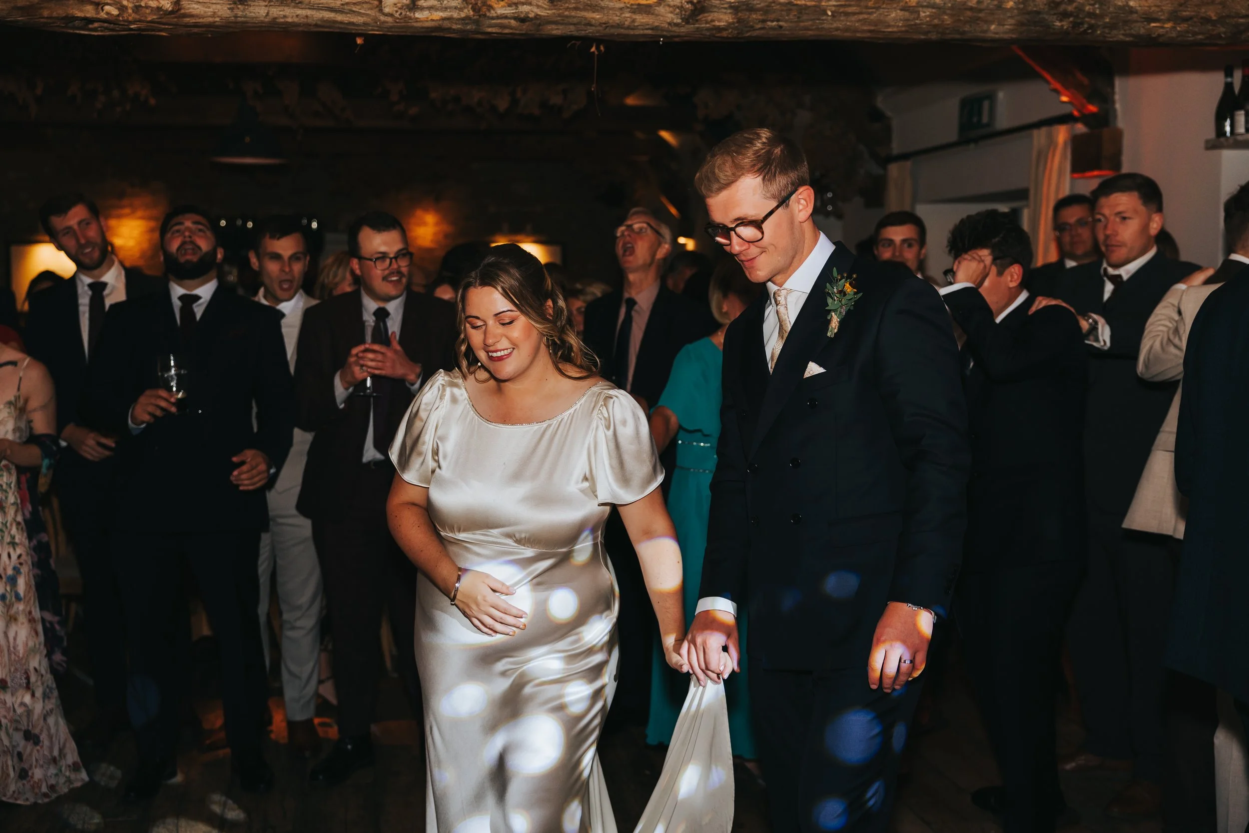 A bride and groom dancing at their wedding reception, surrounded by guests in formal attire, indoors with warm lighting.