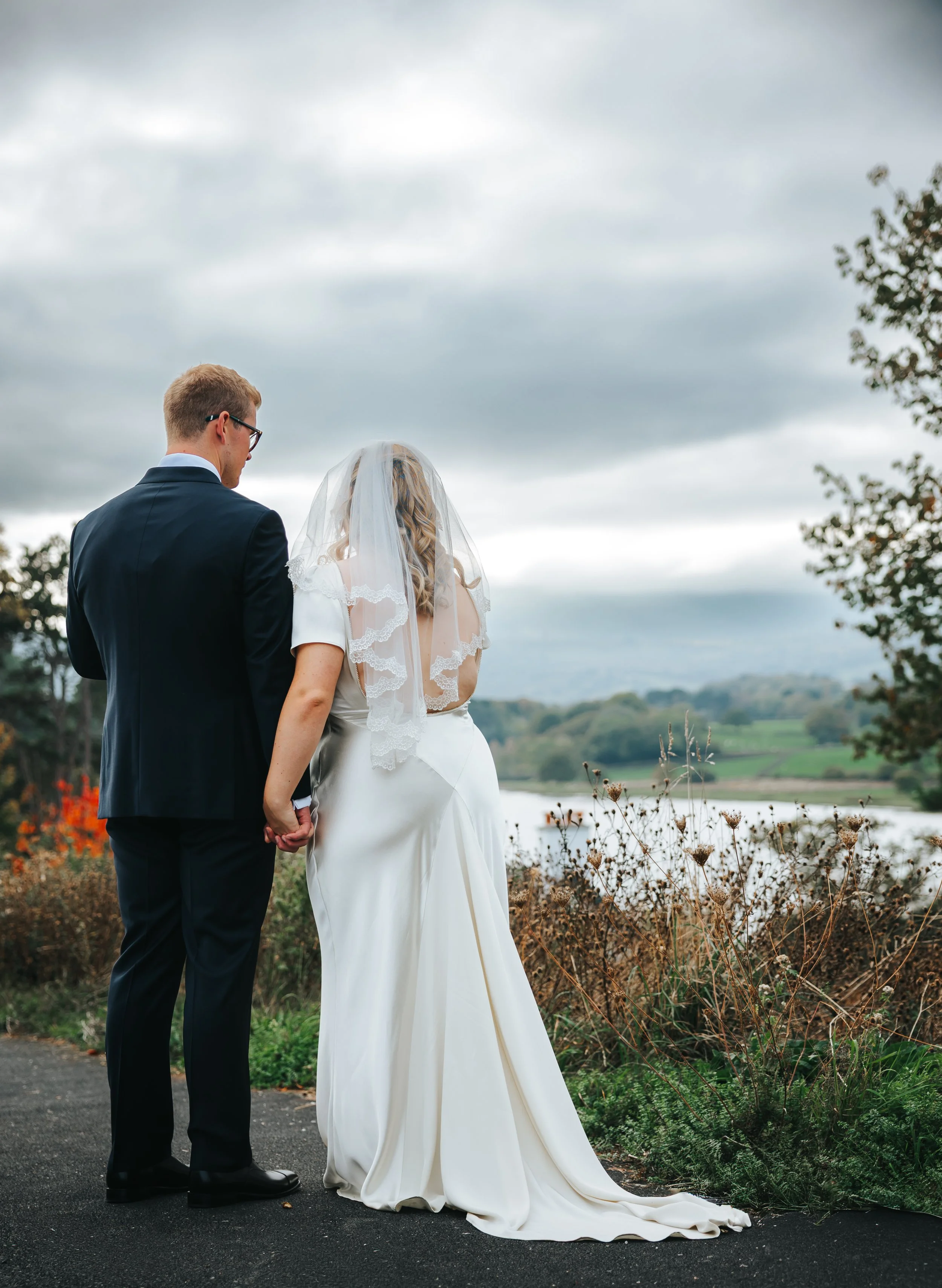 Bride and groom holding hands during their outdoor wedding ceremony by a lake, with cloudy sky in the background.