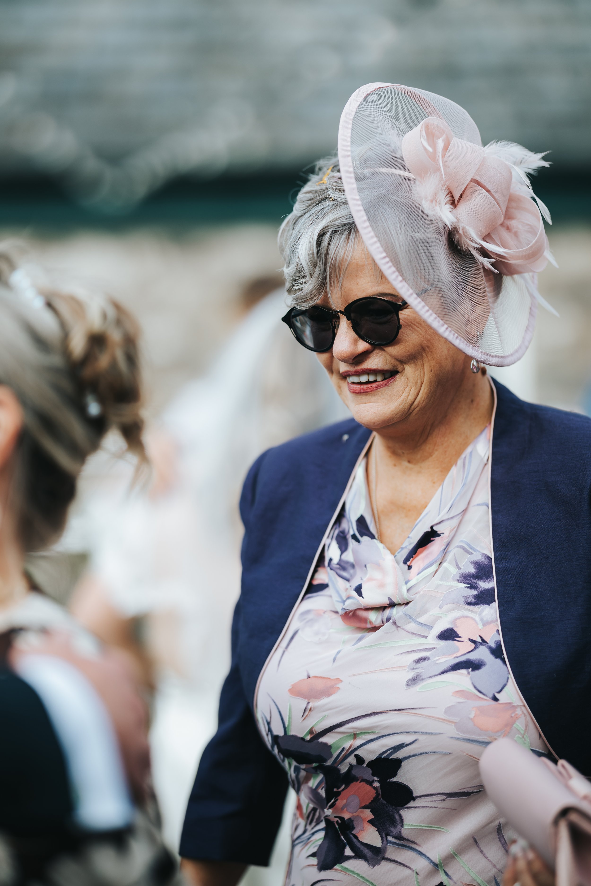 An elderly woman wearing black sunglasses, a pink fascinator with feathers, a floral dress, and a navy blazer, smiling and engaging with a person in front of her at an outdoor event.